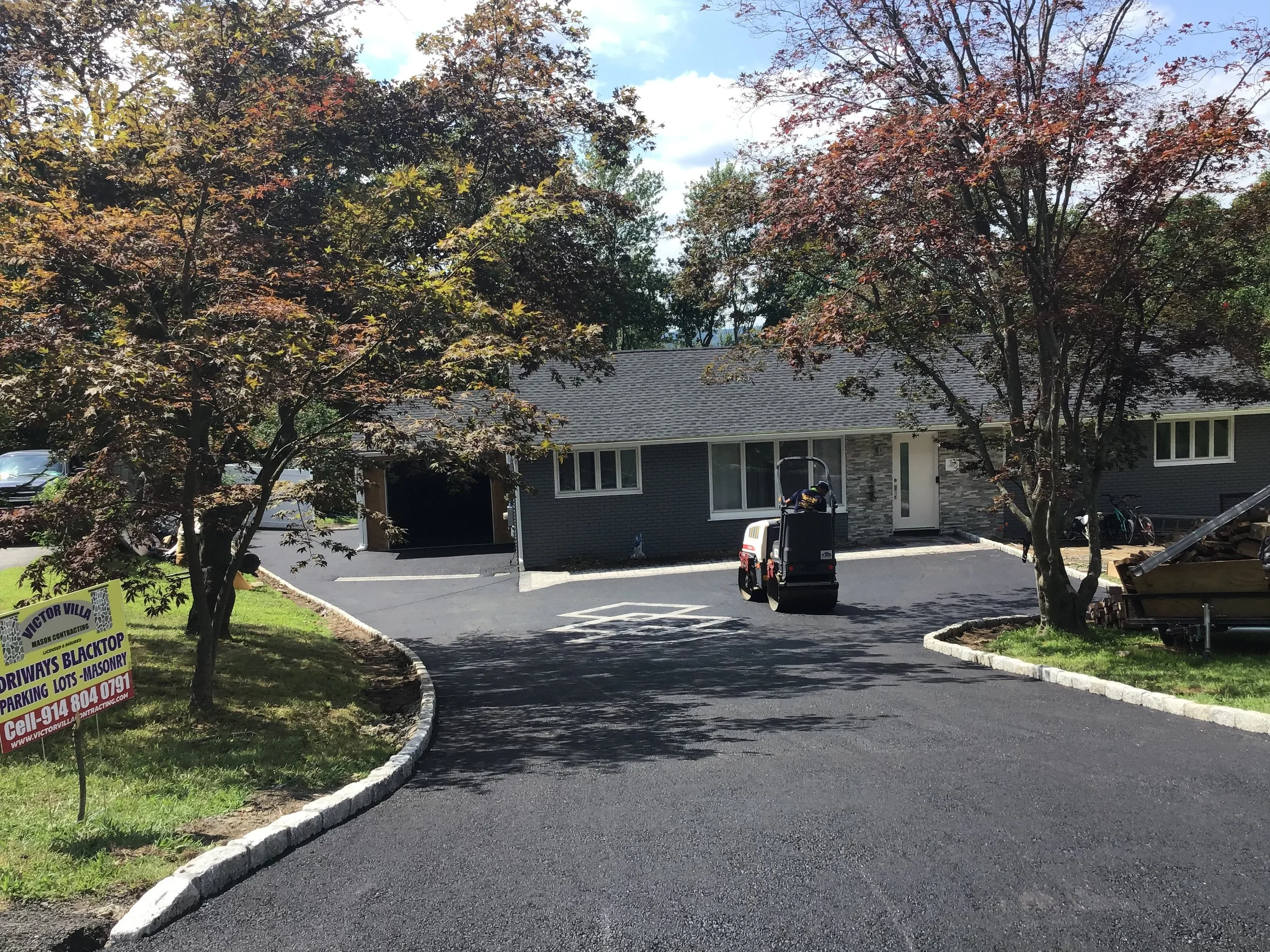 Newly paved asphalt driveway leading to a modern gray house with large window and front door, flanked by two trees with reddish leaves, outdoor lawn sign for driveway blacktop and masonry, and a small vehicle parked in front.