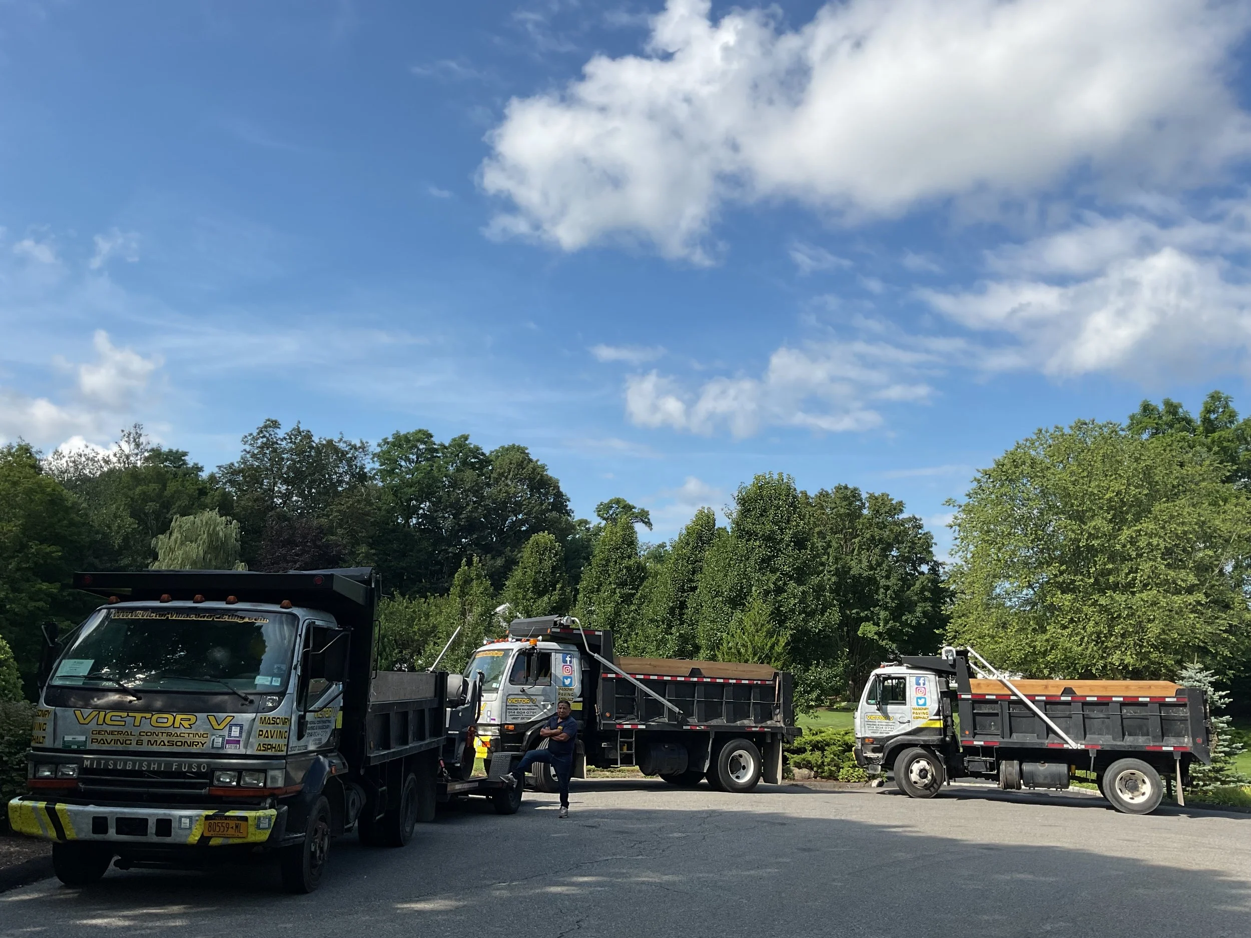 Three construction trucks parked on a paved area with a man sitting on one of the trucks, trees and a partly cloudy blue sky in the background.