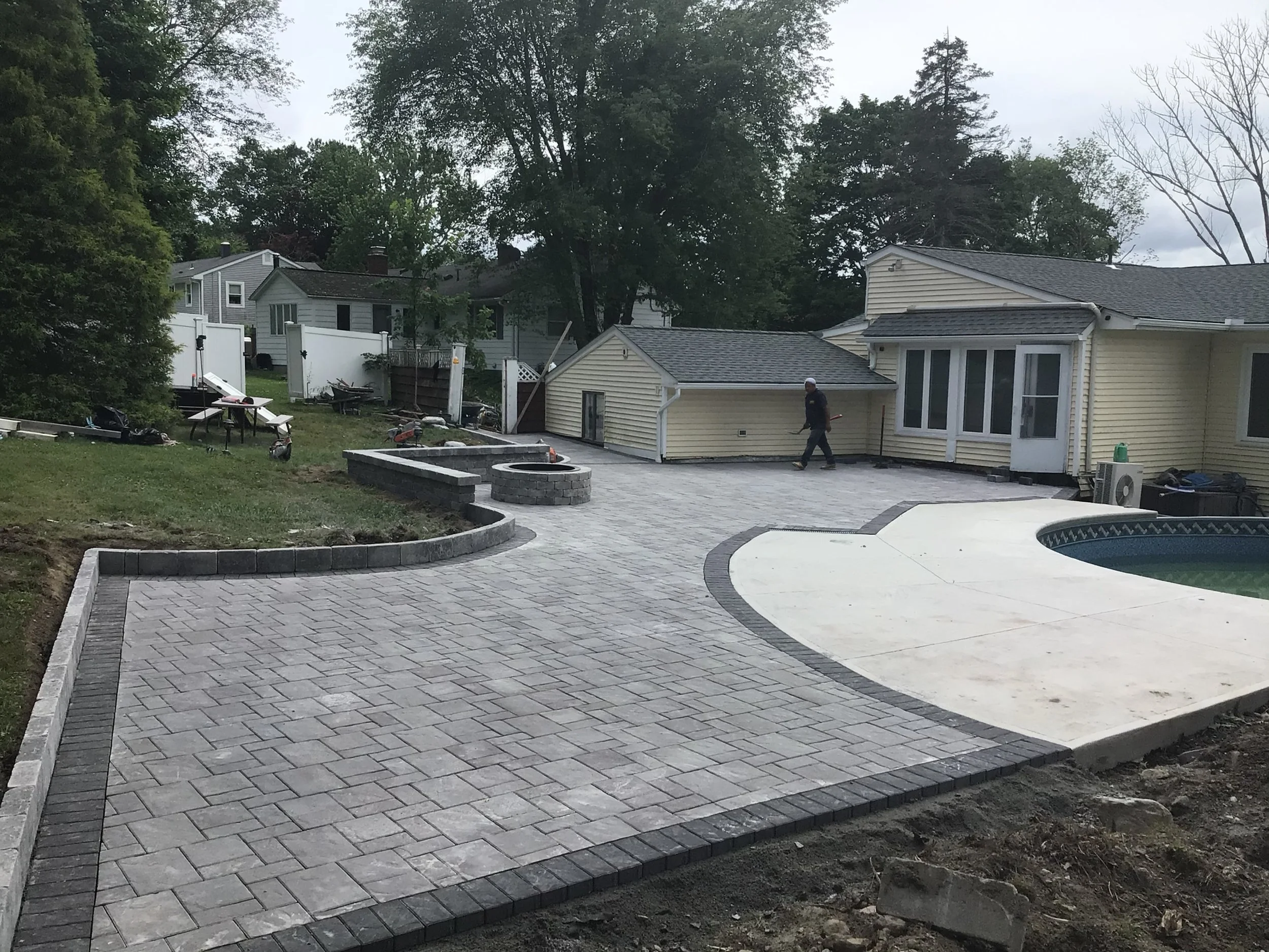 Backyard construction scene with a newly paved patio, a small circular brick fire pit, a swimming pool, and a worker walking across the patio near a house with yellow siding.