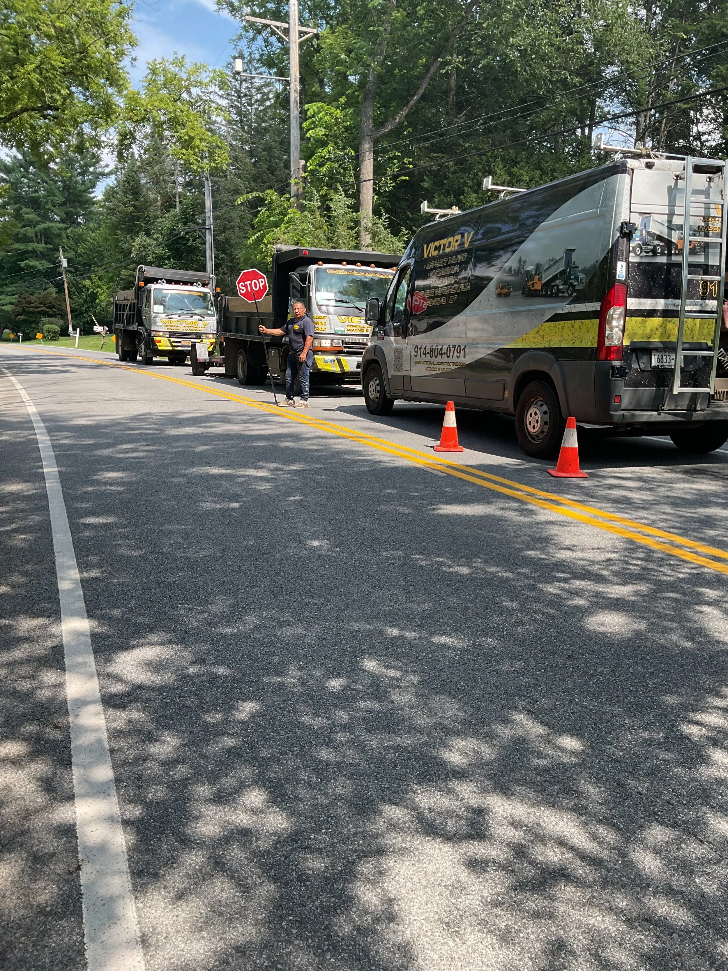 Several vehicles including a van and two trucks are parked on a road with orange cones. A worker is holding a stop sign in the middle of the scene, and there are trees and power lines in the background.