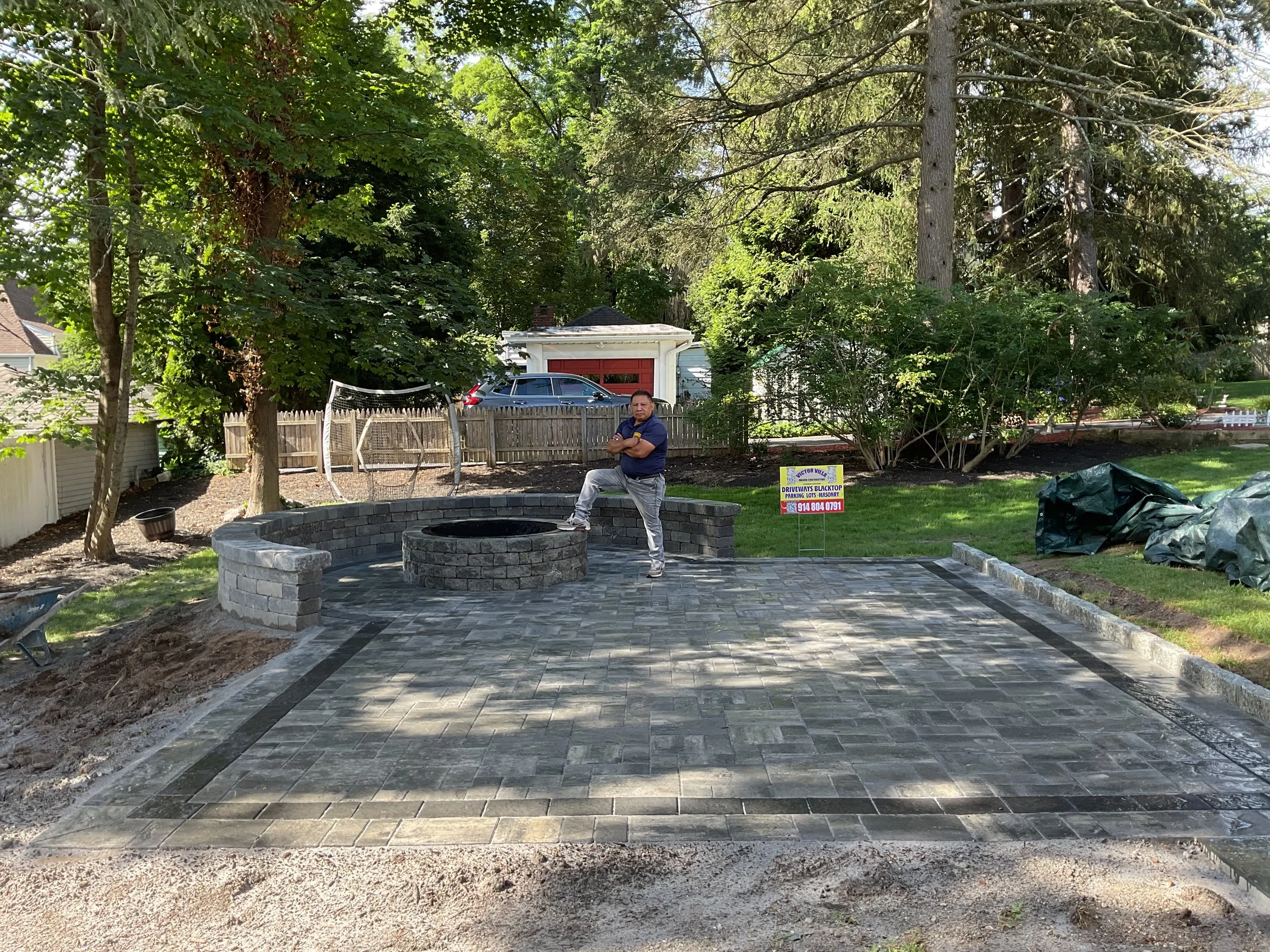 A man standing on a newly constructed brick patio with a fire pit in a backyard, trees and a fence in the background, and signs indicating construction work.