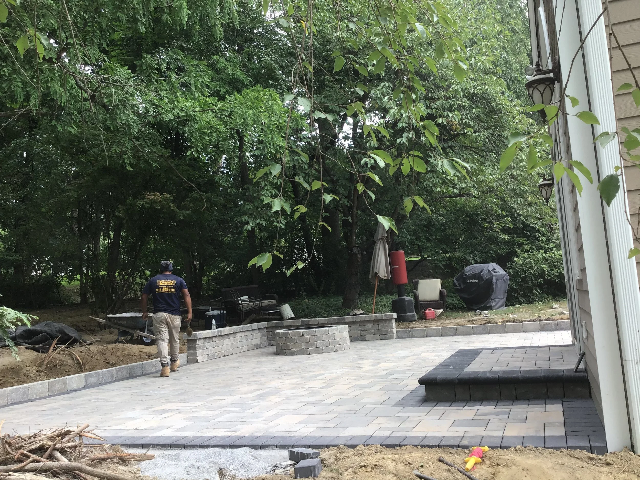 A man working on a backyard patio construction with pavers, a brick wall, and outdoor furniture, surrounded by trees and ongoing landscaping.