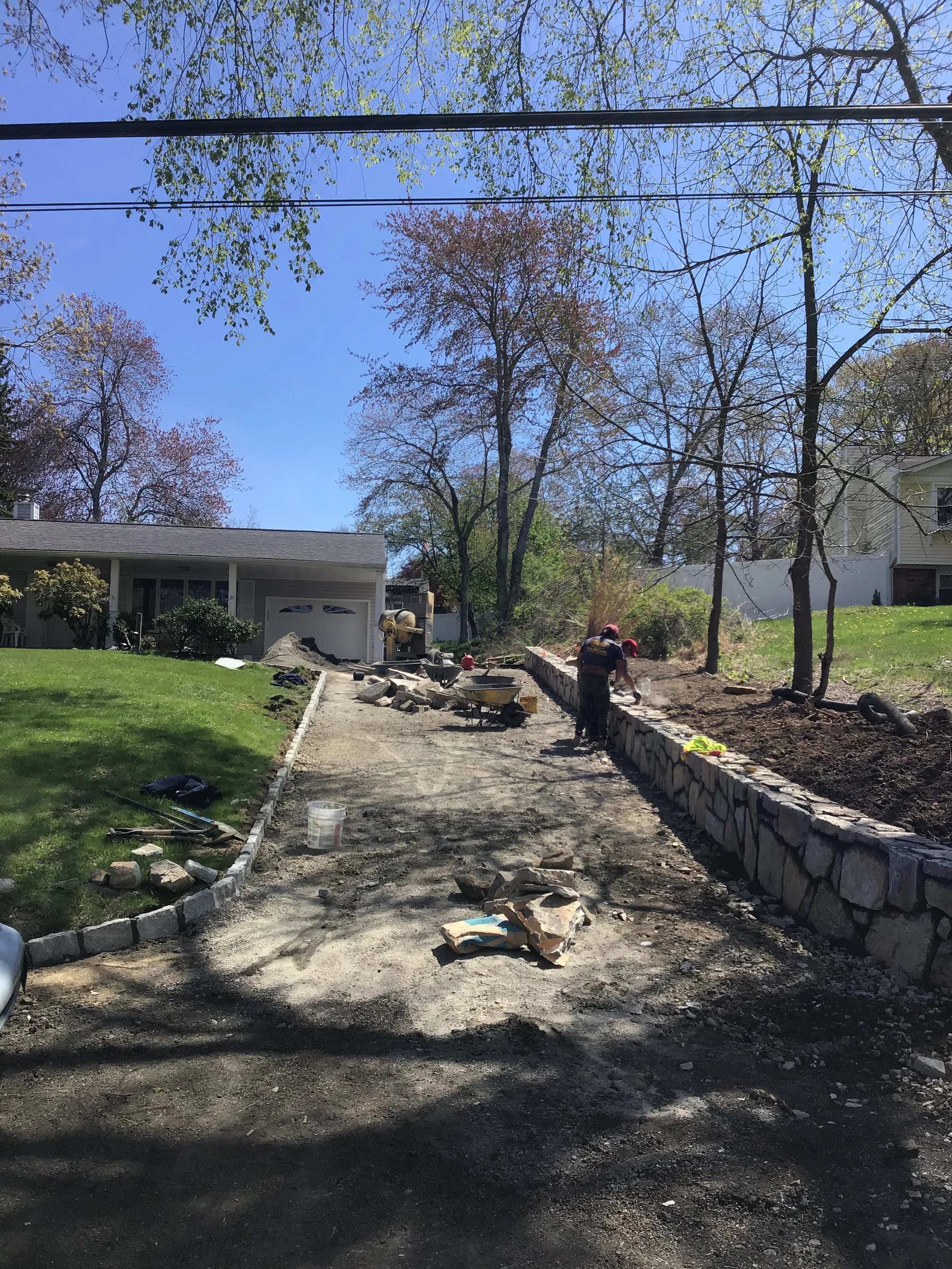 Construction workers building a stone wall along a driveway in a suburban neighborhood on a sunny day with trees and houses in the background.