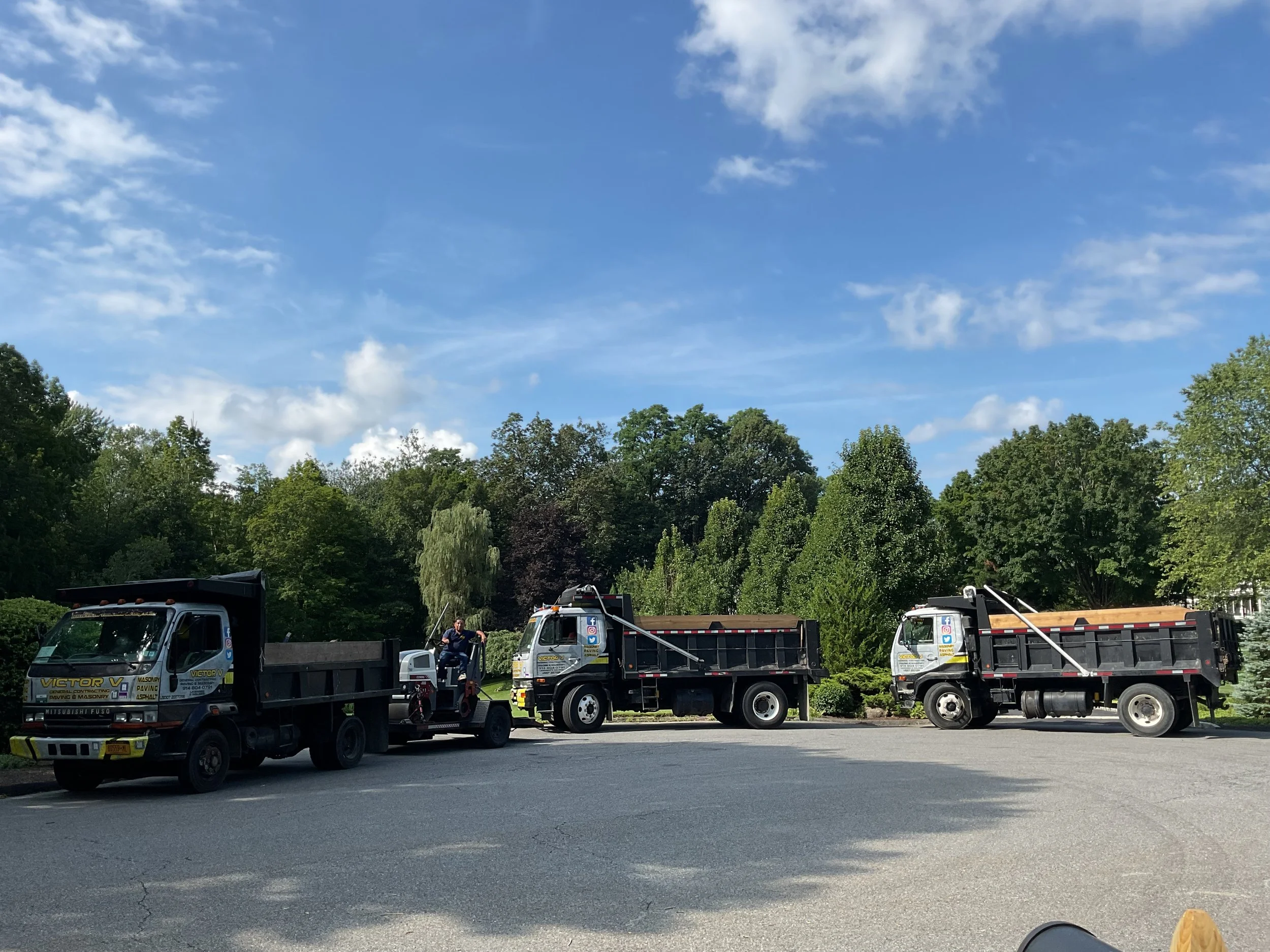 Three trucks parked on the side of a street with trees and blue sky in the background.