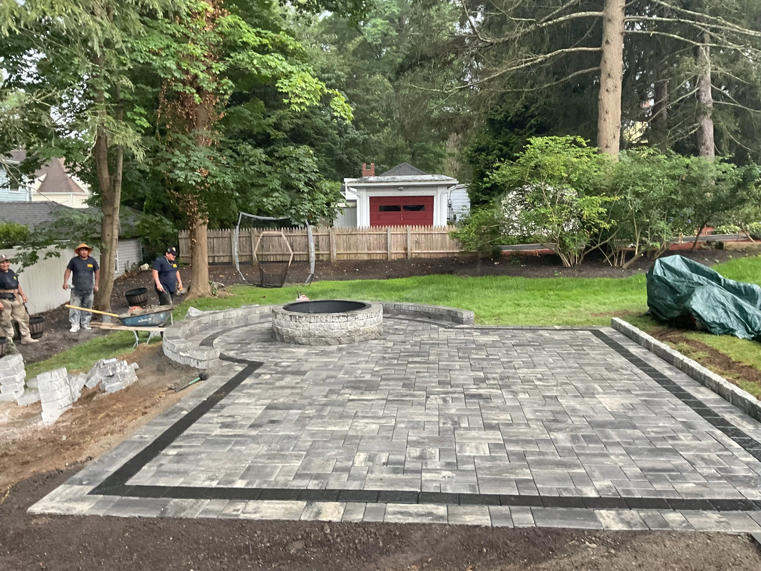 Workers installing a paved patio in a backyard with trees, a wooden fence, and a small shed in the background.