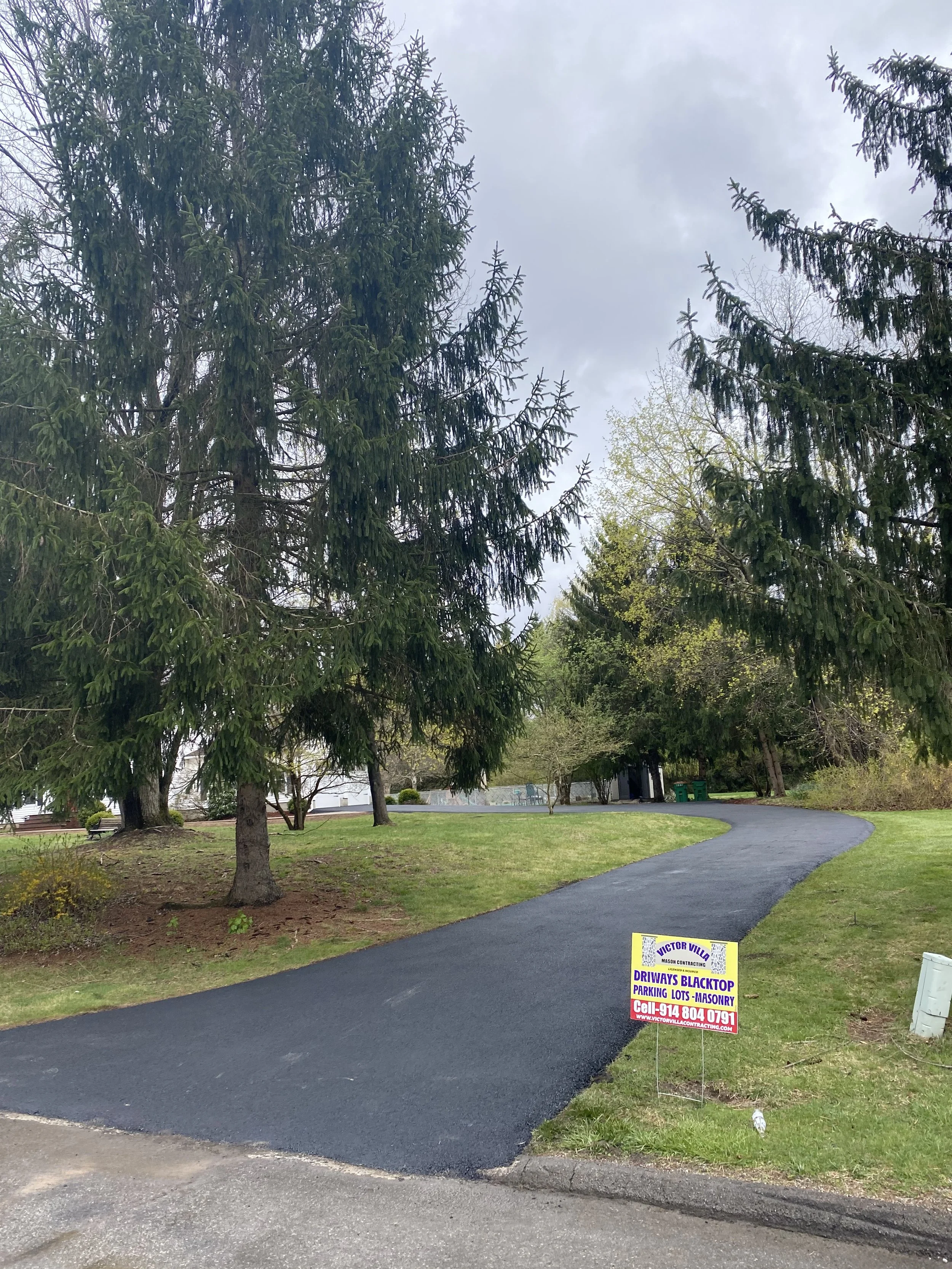 A paved driveway or pathway in a residential or park area surrounded by green grass and tall trees under a cloudy sky, with a sign at the entrance advertising driveway blacktop paving services.