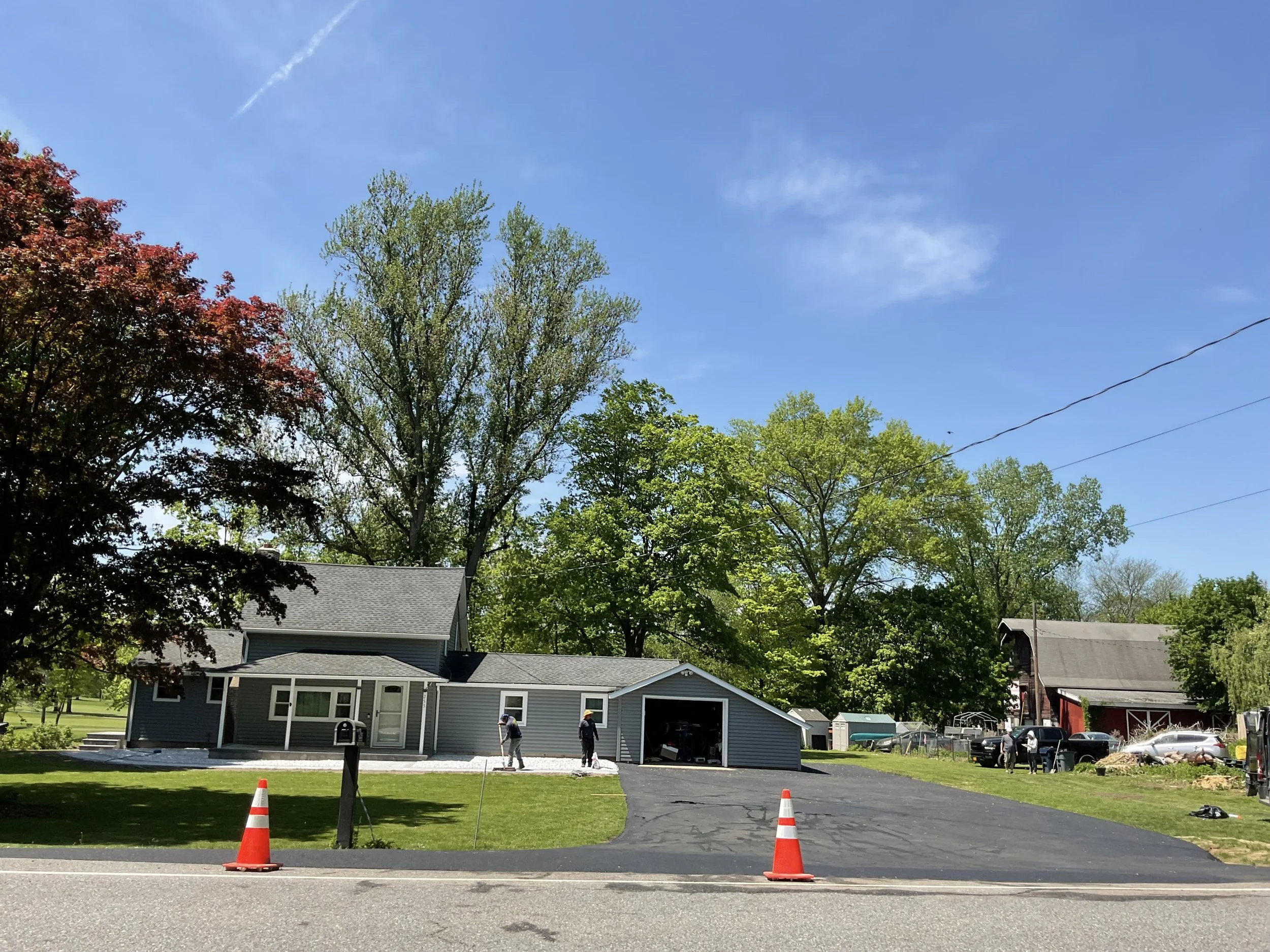 Residential house with renovation workers, orange traffic cones, trees, blue sky, and a barn in the background