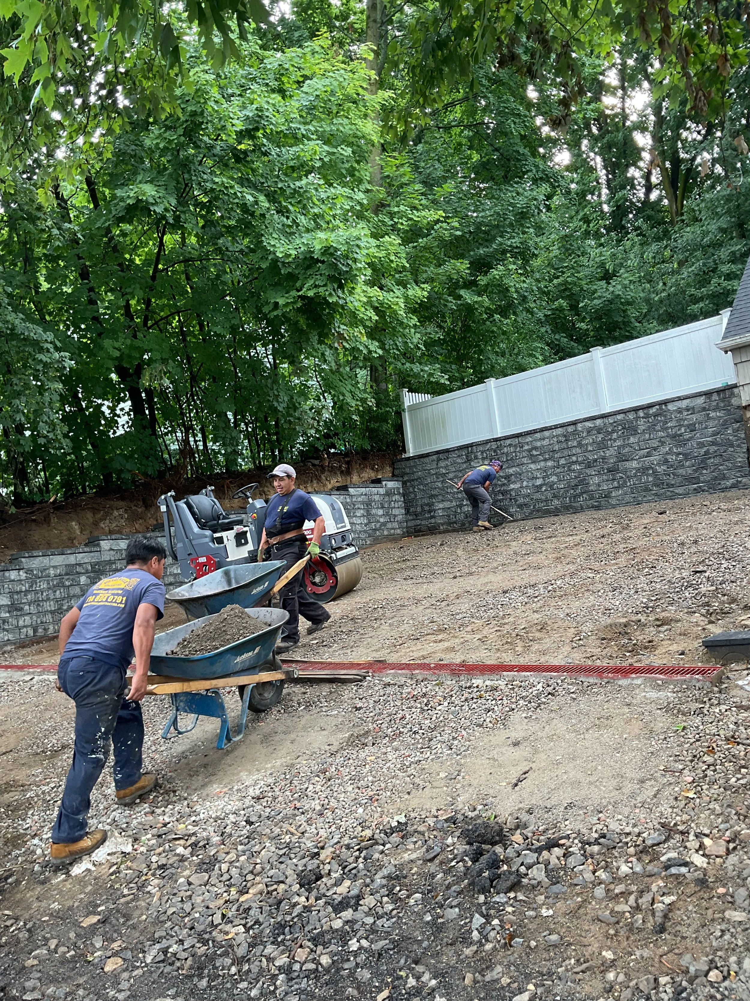 Three workers are laying down a gravel path on a sloped yard. One worker is pushing a wheelbarrow full of gravel, another is raking gravel on the ground, and the third is leveling the gravel near a retaining wall with a compactor machine nearby. Ther