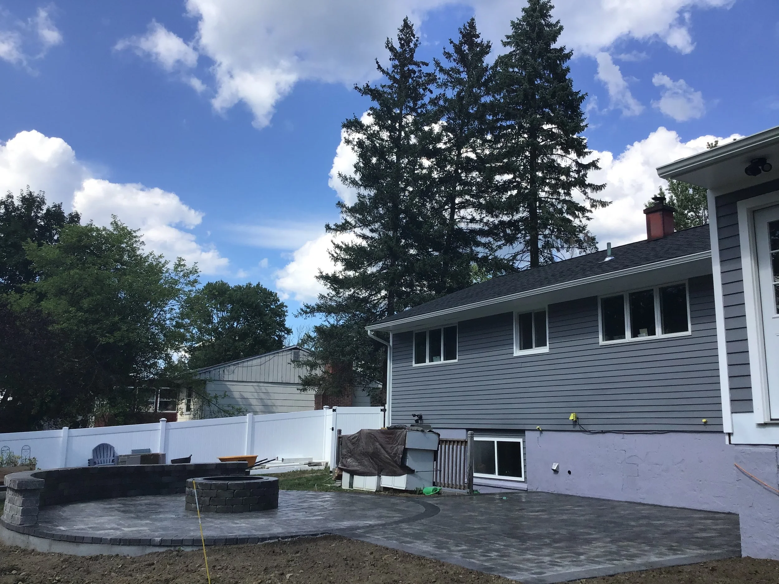 Backyard with newly paved patio area, a white fence, trees, and a gray house with multiple windows.