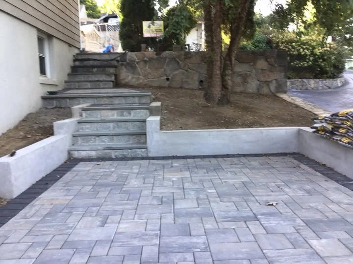 Newly paved driveway with stone steps leading to a higher level, bordered by white concrete walls, with dirt and trees in the background.