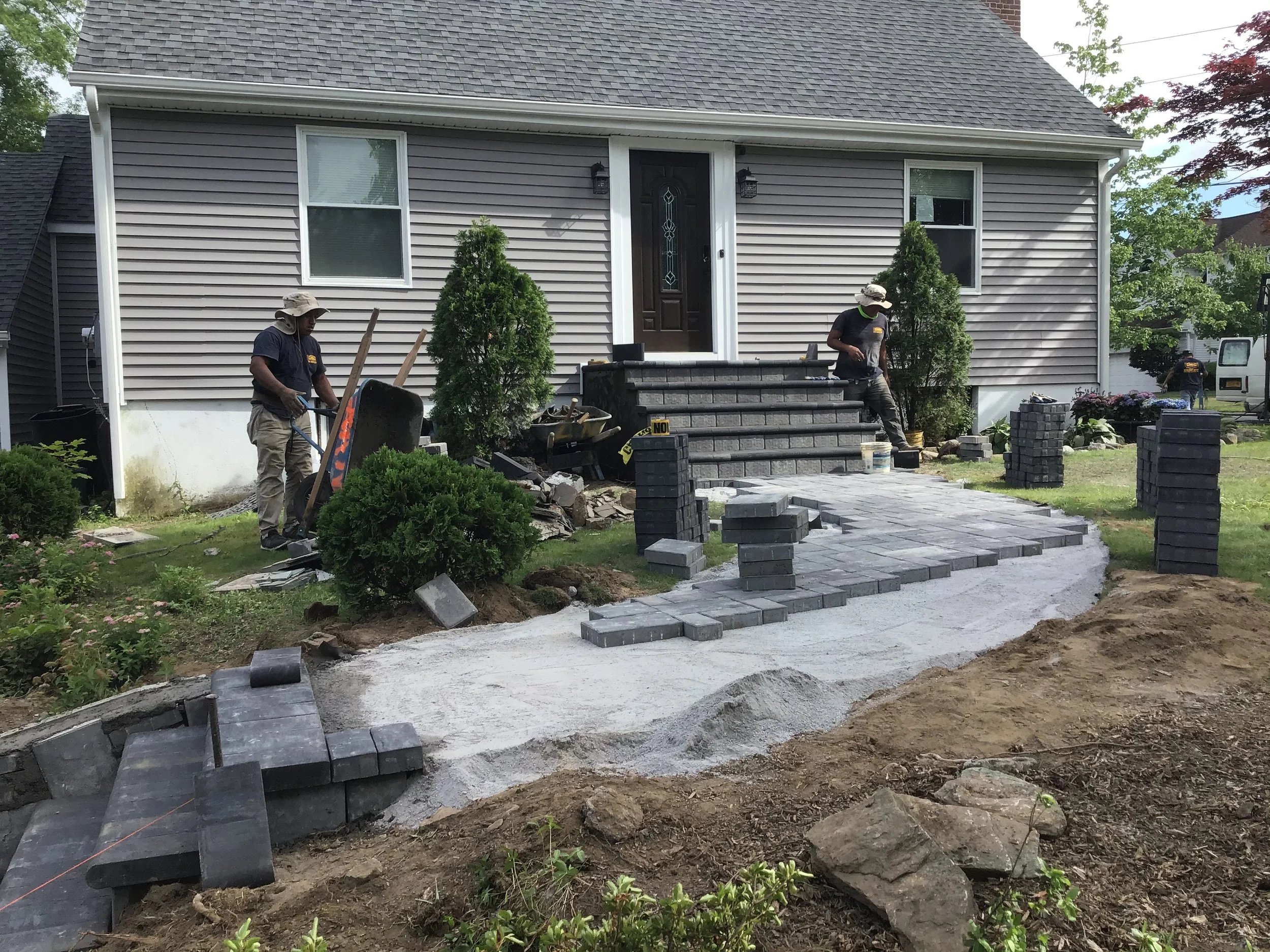 Workers installing gray brick pavers and constructing a new pathway in front of a house during landscaping project.