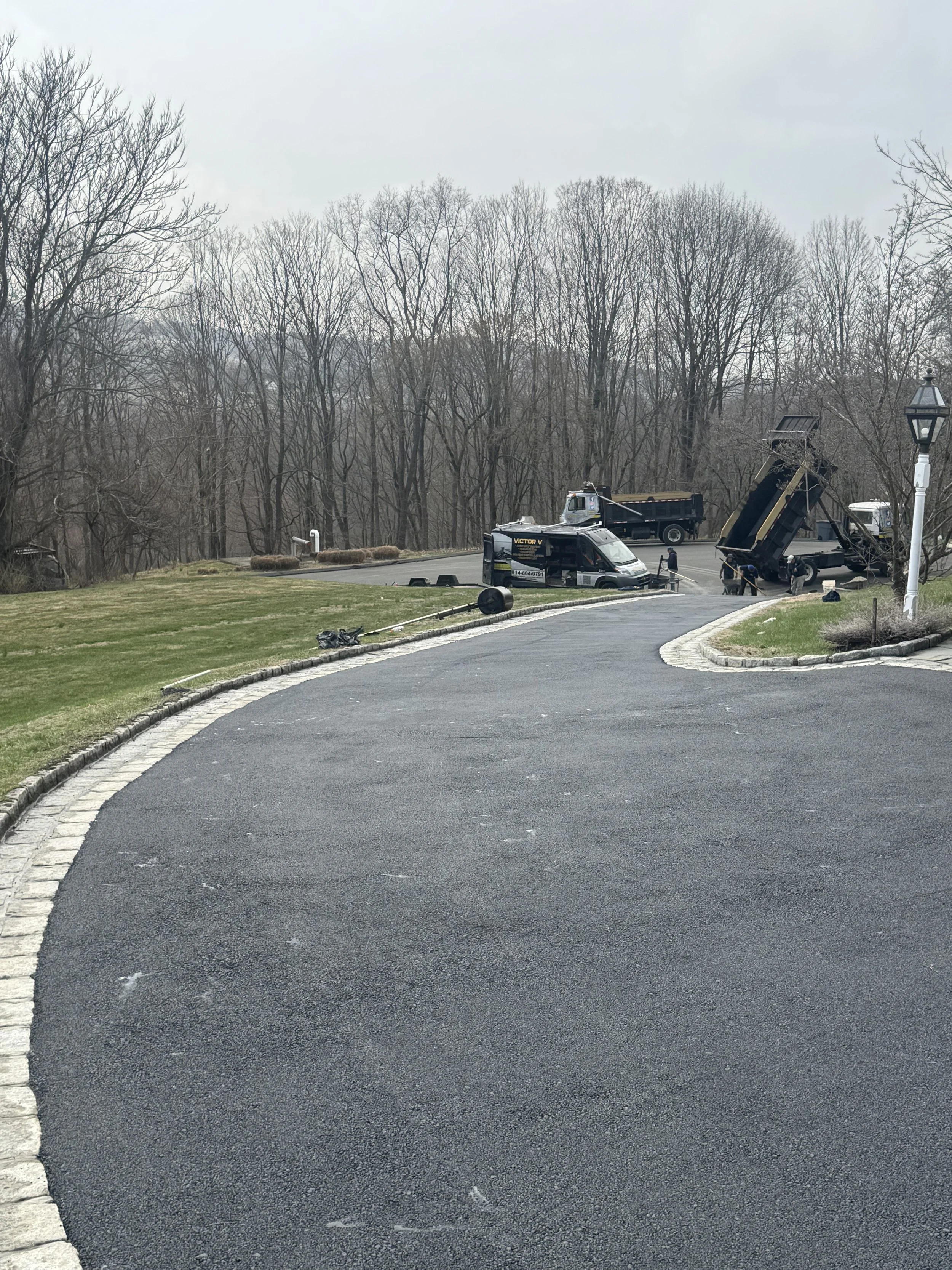 A paved driveway with a curved edge leading to a yard with green grass and leafless trees in the background. There is construction or maintenance equipment and vehicles parked at the end of the driveway, including a truck with a crane and a van. Work