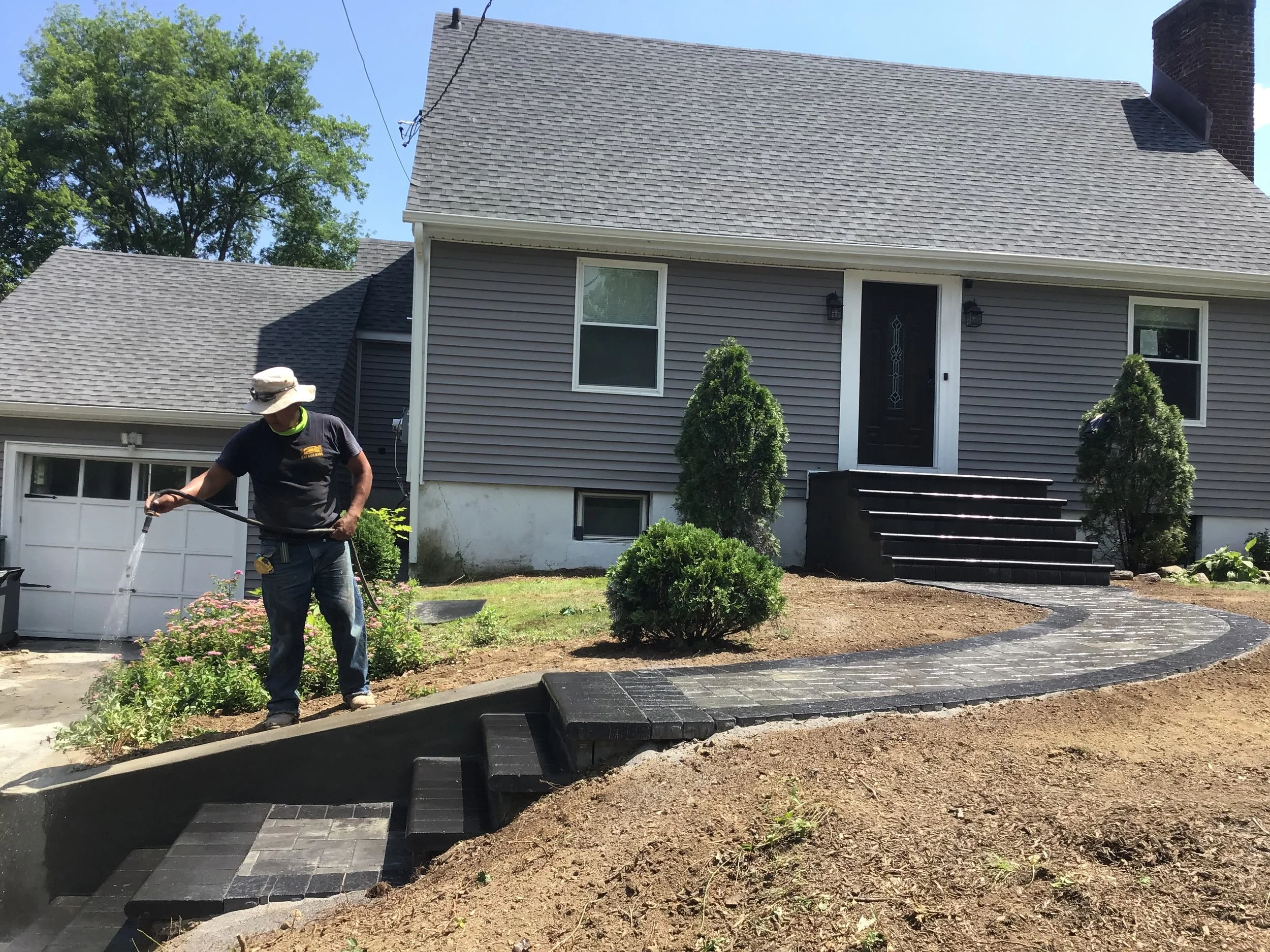A person stands on a freshly landscaped backyard near a house, watering newly laid black stone pathway and steps.