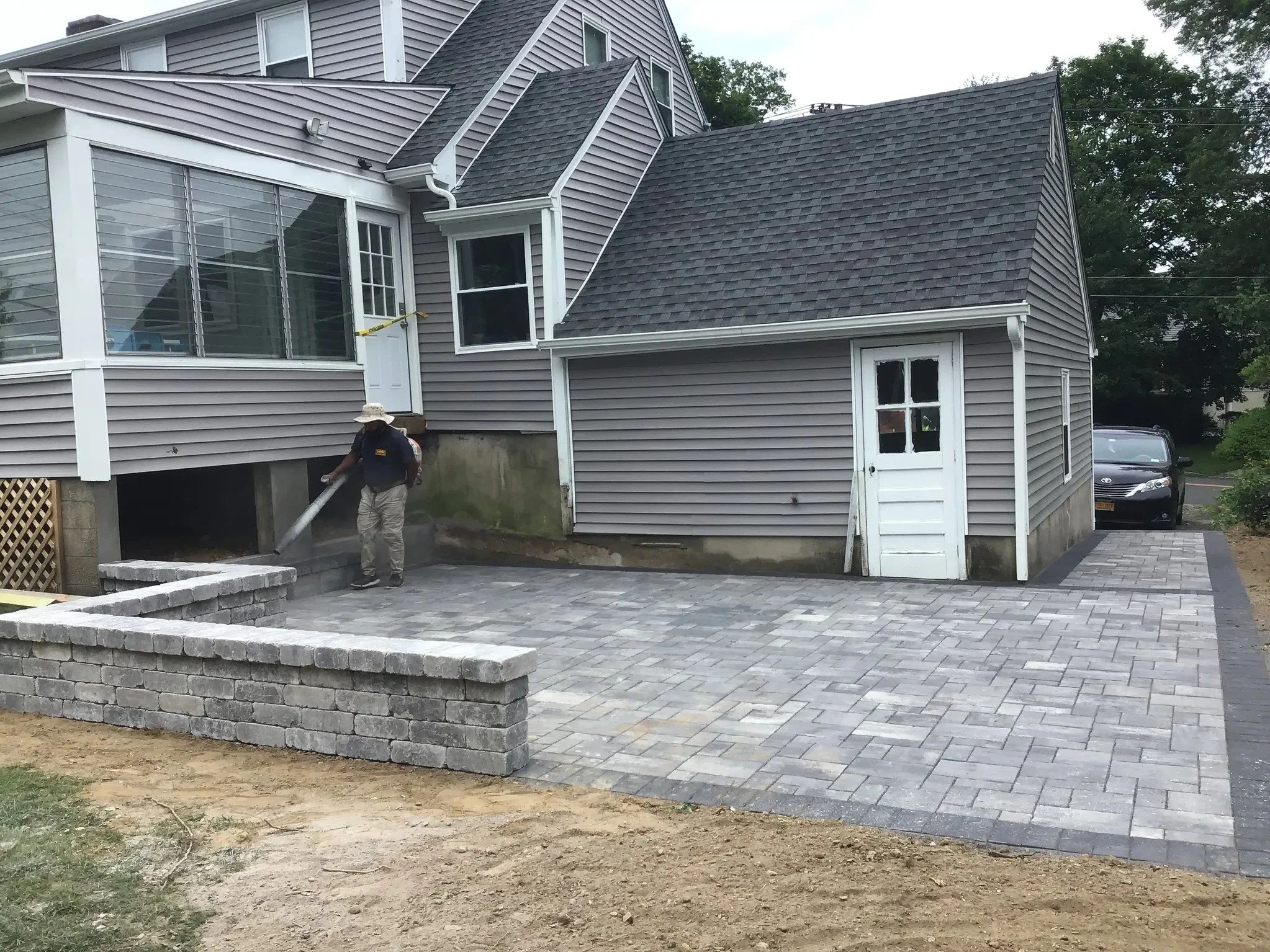 Person paving a backyard with gray stone bricks, with a gray house and a parked car in the background.