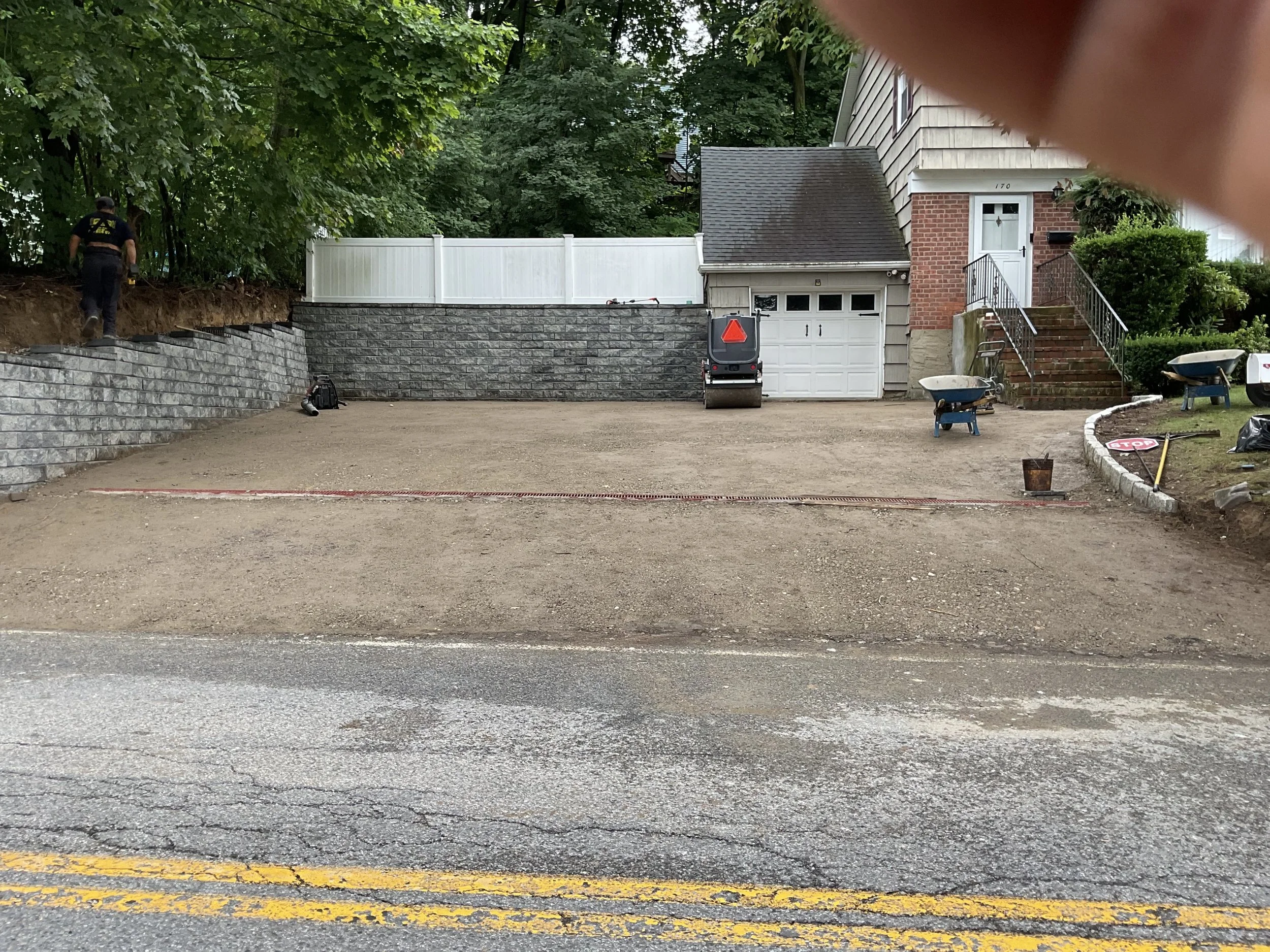 A residential driveway during construction with a worker, construction equipment, and tools, and a wheelbarrow blanketed with dirt.