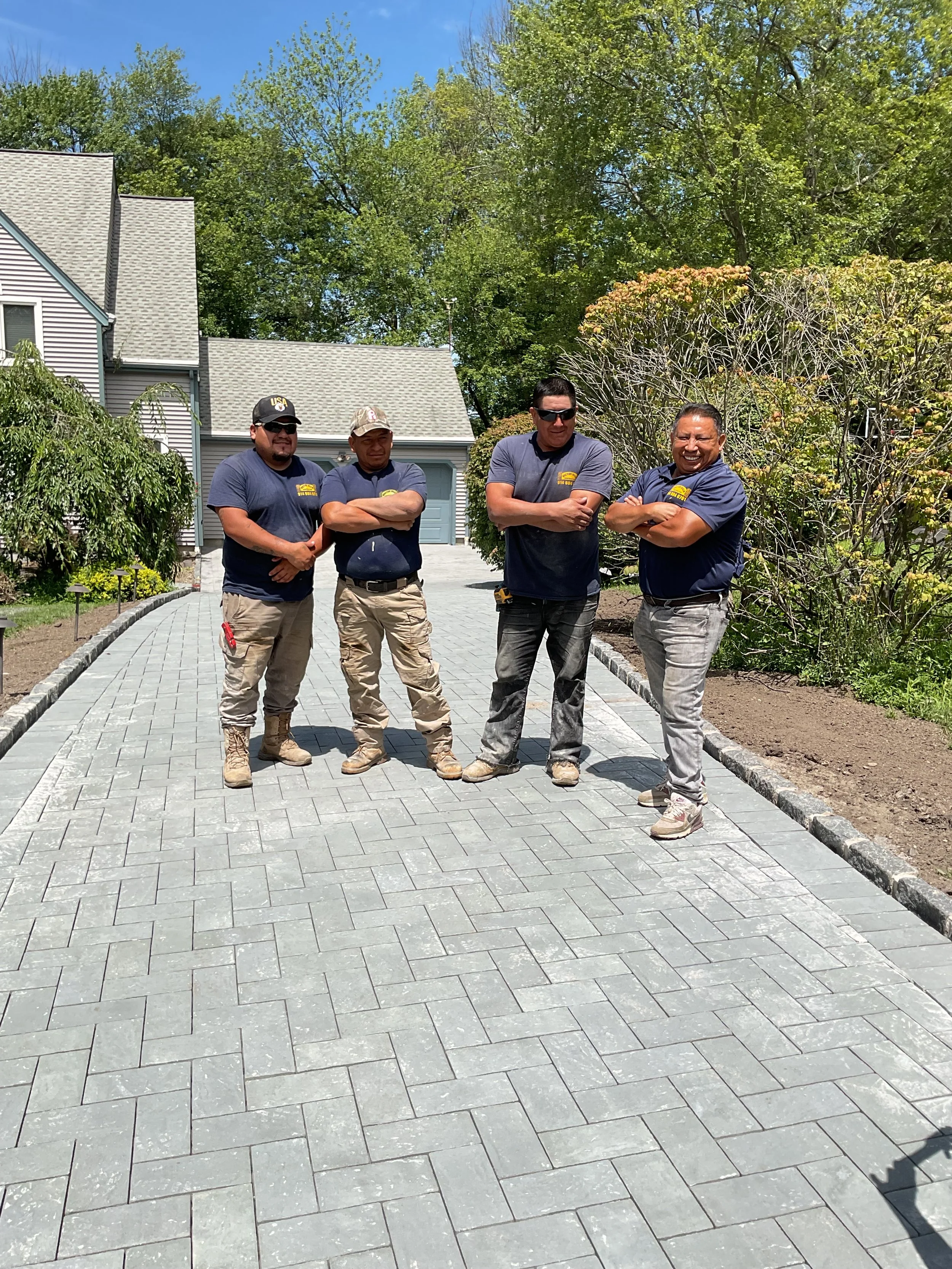 Four workers in navy shirts and work pants standing on a newly paved stone driveway in front of a house, with trees and bushes in the background.