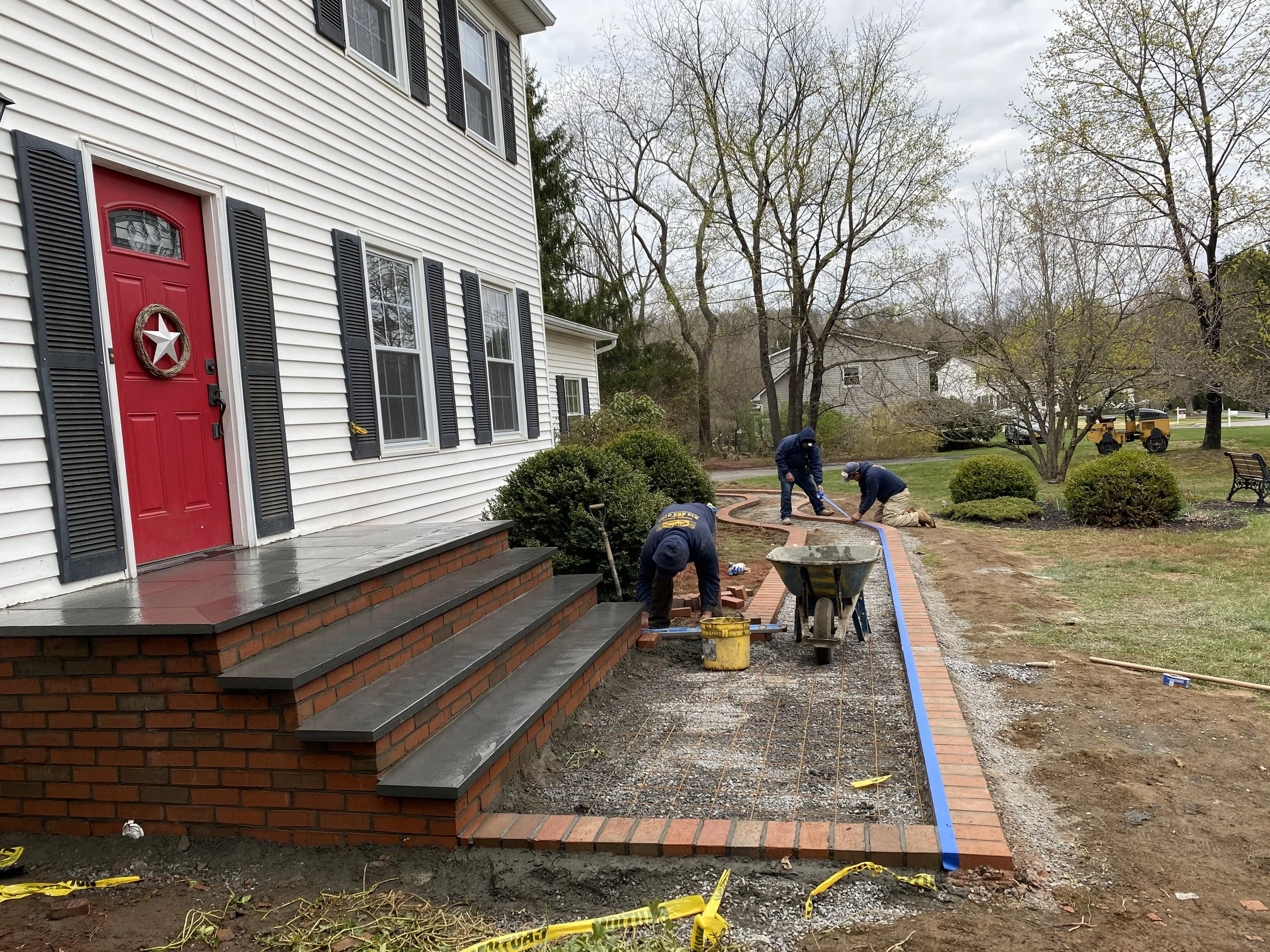 Workers constructing a brick walkway outside a white house with red door and black shutters, on a cloudy day.