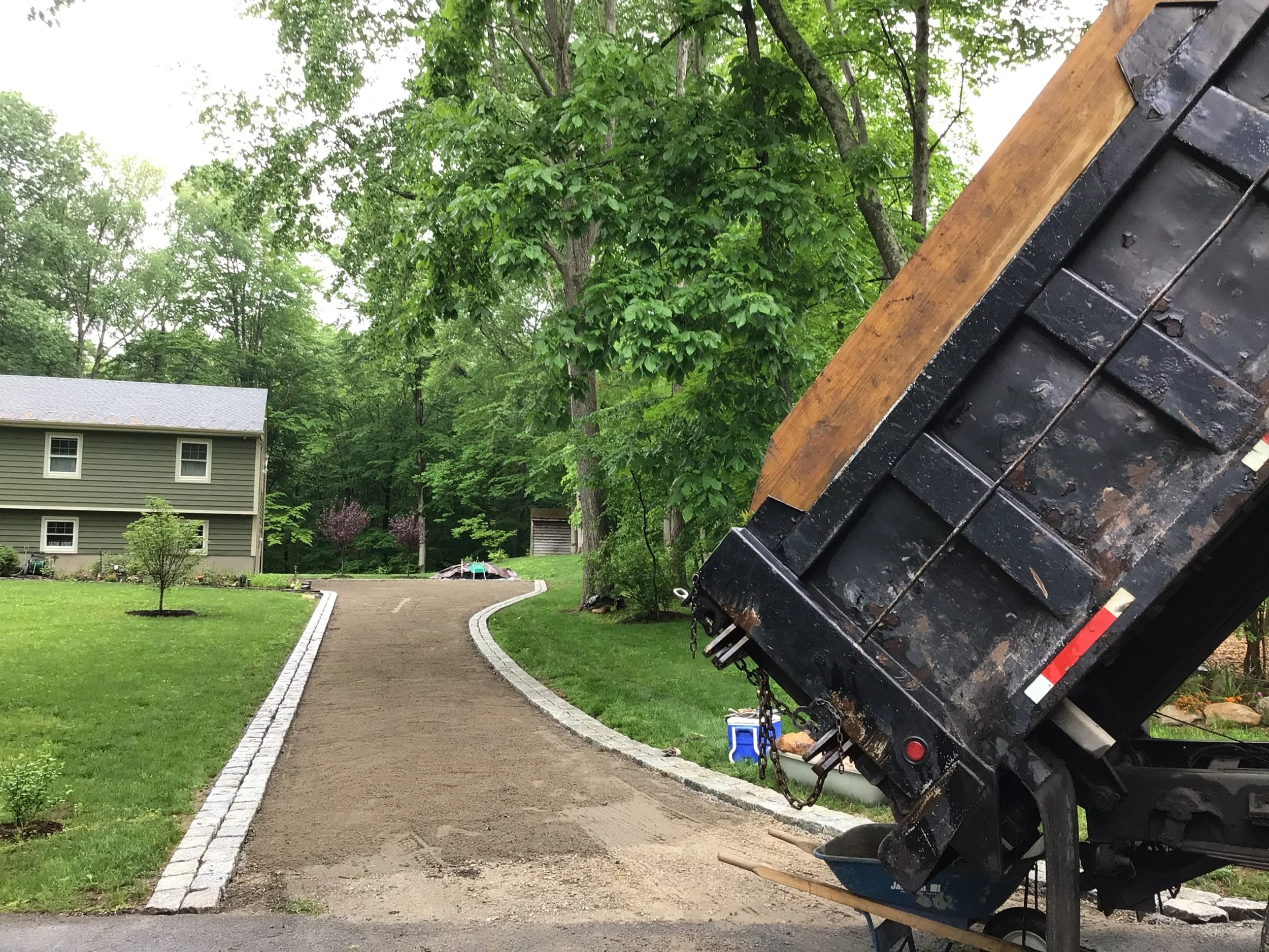 A driveway under construction with a dump truck unloading gravel or dirt, surrounded by green grass, trees, and a house with siding.