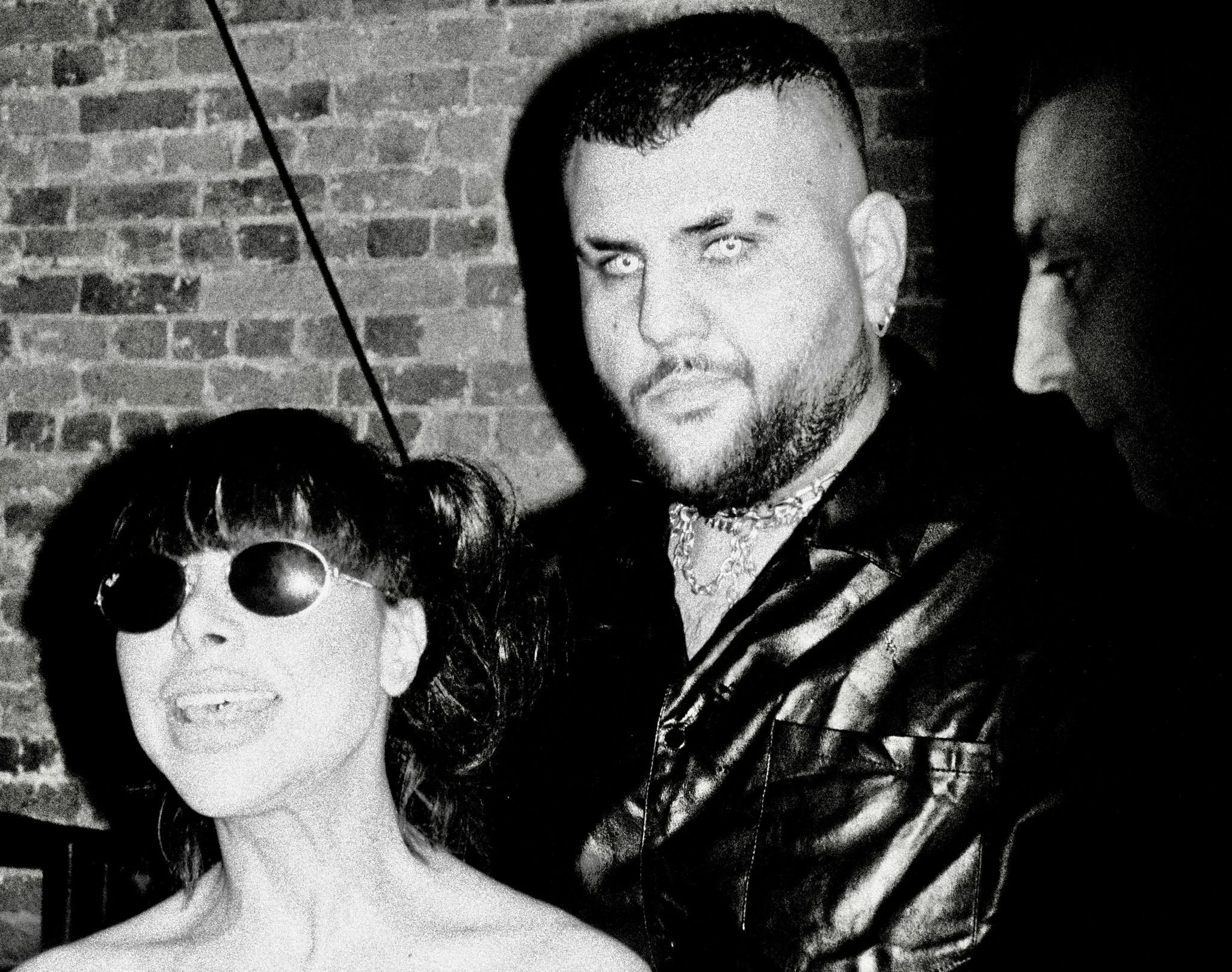 A black-and-white photo of three people: a woman with short, curly hair and round sunglasses smiling widely, a man with short dark hair, a beard, and intense eyes looking at the camera, and another person with short hair visible on the right side, ag