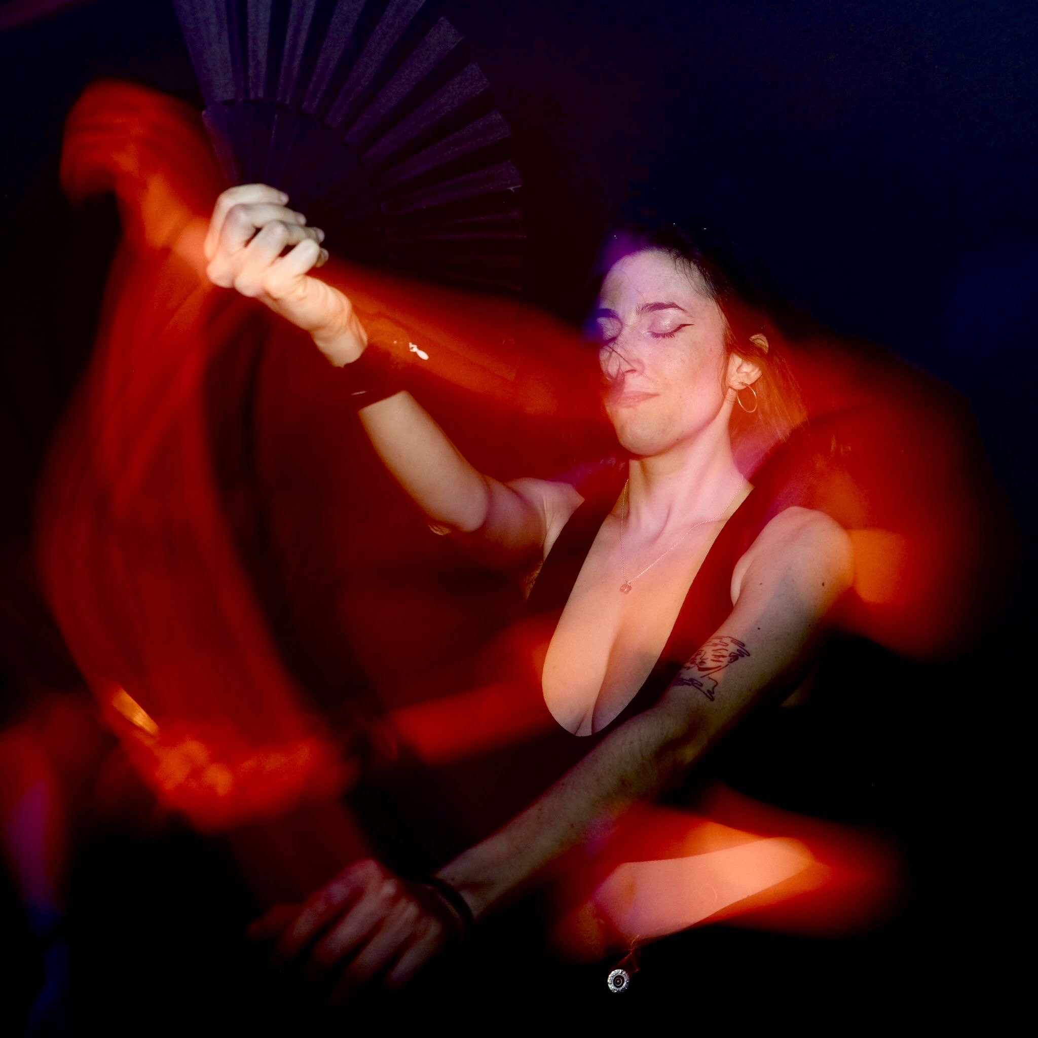 A woman with short dark hair and tattoos on her arm, holding a fan, dancing in a dark environment with red and orange light streaks.