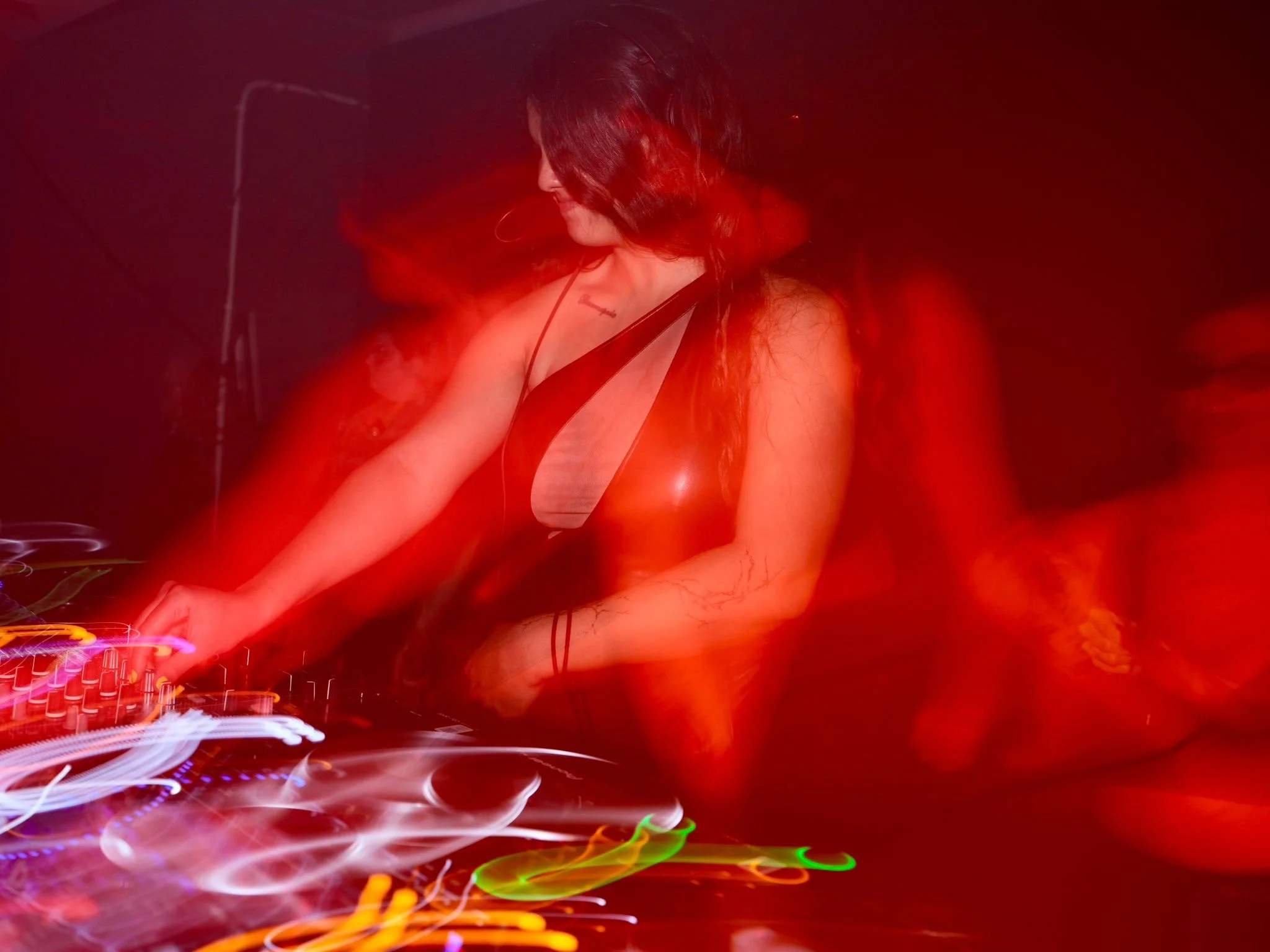 A woman DJ wearing a revealing black top with a cutout design, mixing music at a club with colorful lighting effects.