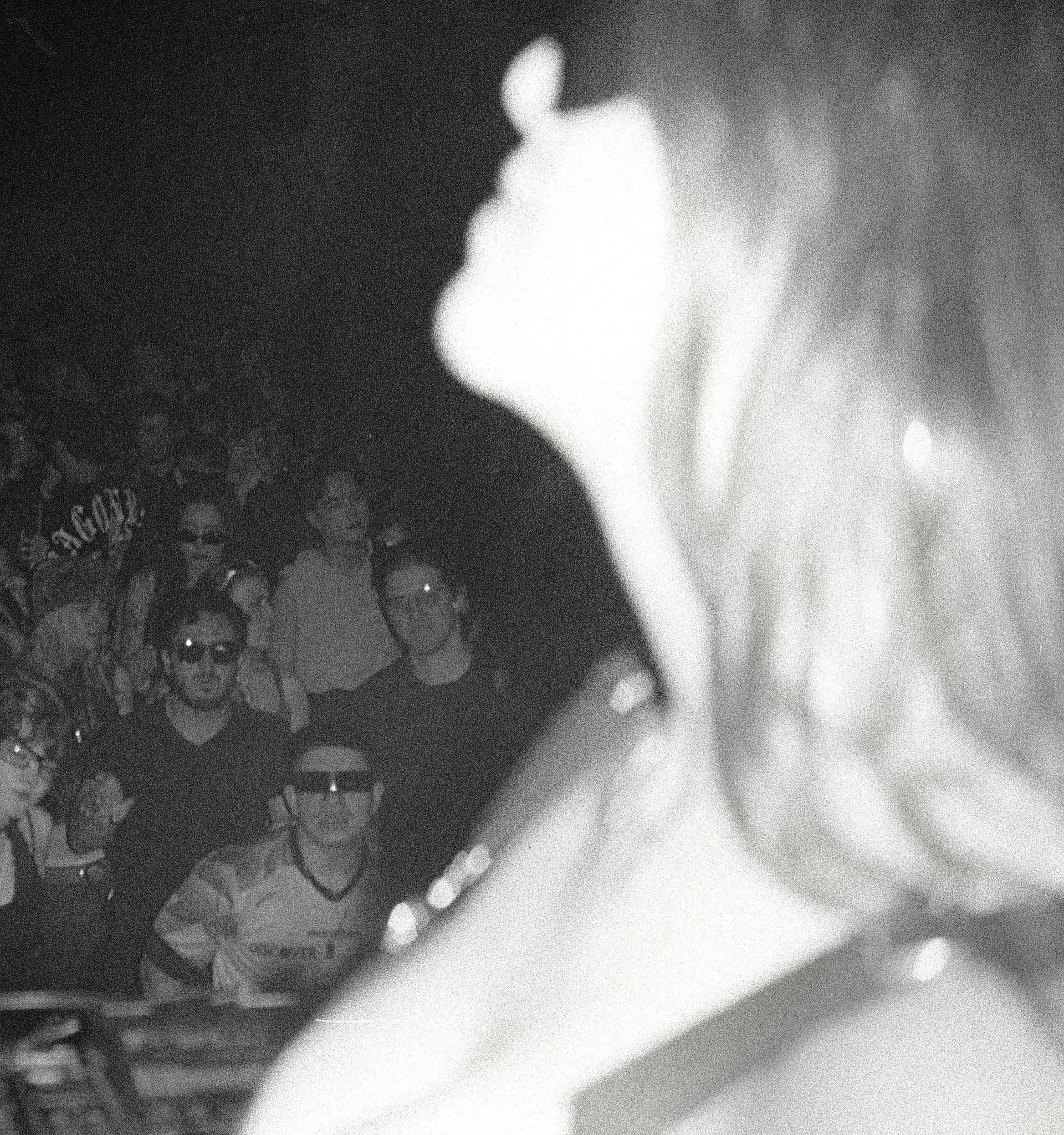 A black-and-white photo taken through a reflective surface shows a crowd of people, some wearing glasses, in the background. The reflection displays a partially visible hand or arm in the foreground.