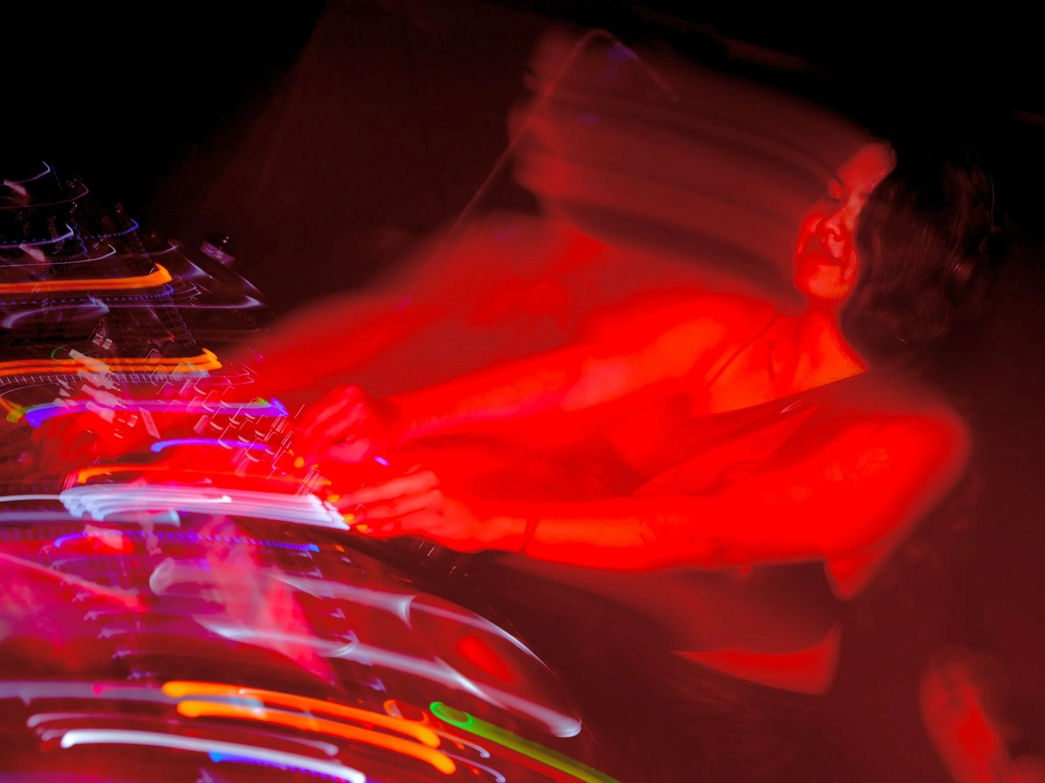 A female DJ with dark hair and headphones, mixing music with colorful light trails on her equipment at a nighttime event.