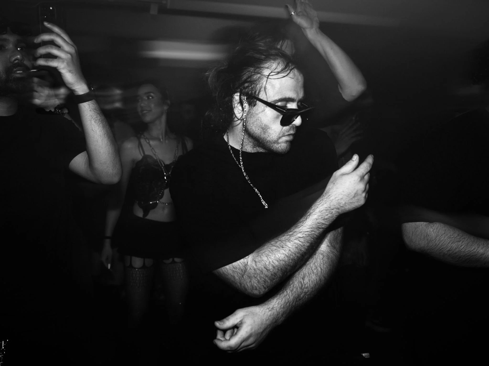 People dancing at a party, with one man in the foreground wearing sunglasses and headphones, looking at his phone, and others dancing in the background.