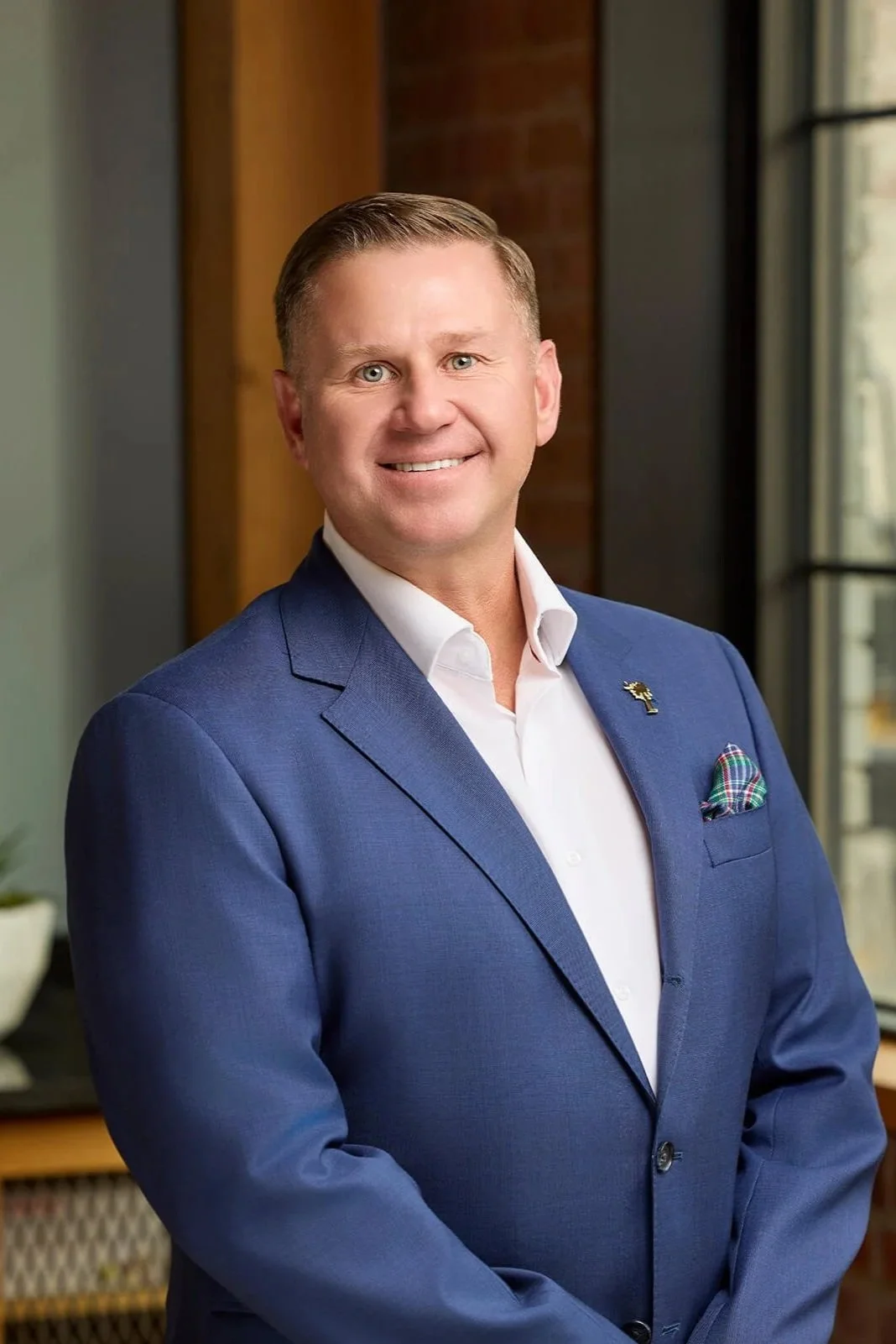 A man in a blue suit with a white shirt and a plaid pocket square, standing indoors by a window with bridges and buildings outside.
