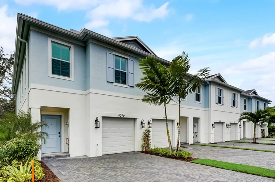 A row of modern two-story townhouses with white and light blue exterior, gray garage doors, palm trees, and well-maintained landscaping under a partly cloudy sky.