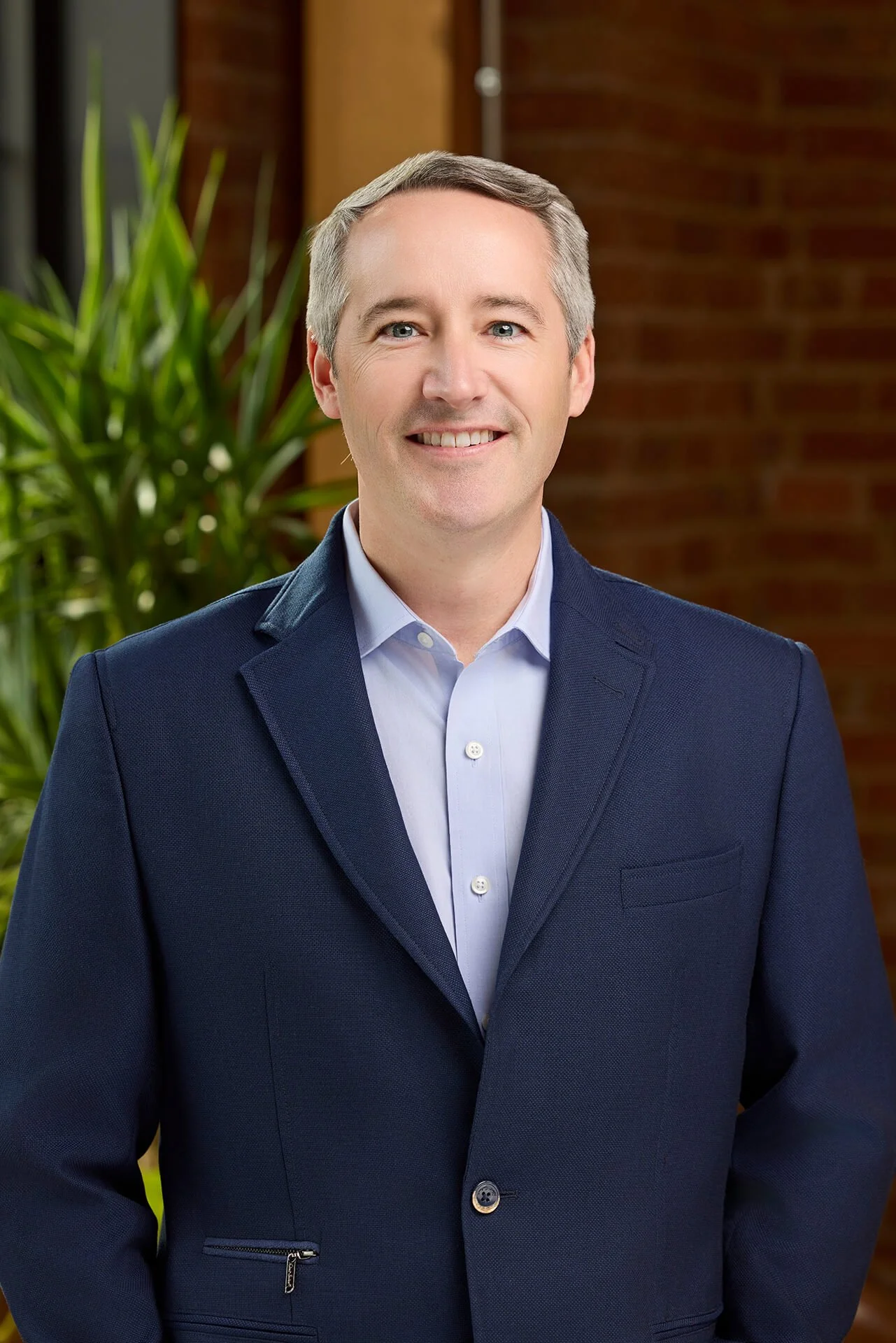 A smiling man in a dark blue blazer and light blue shirt standing in front of a brick wall and green plant.
