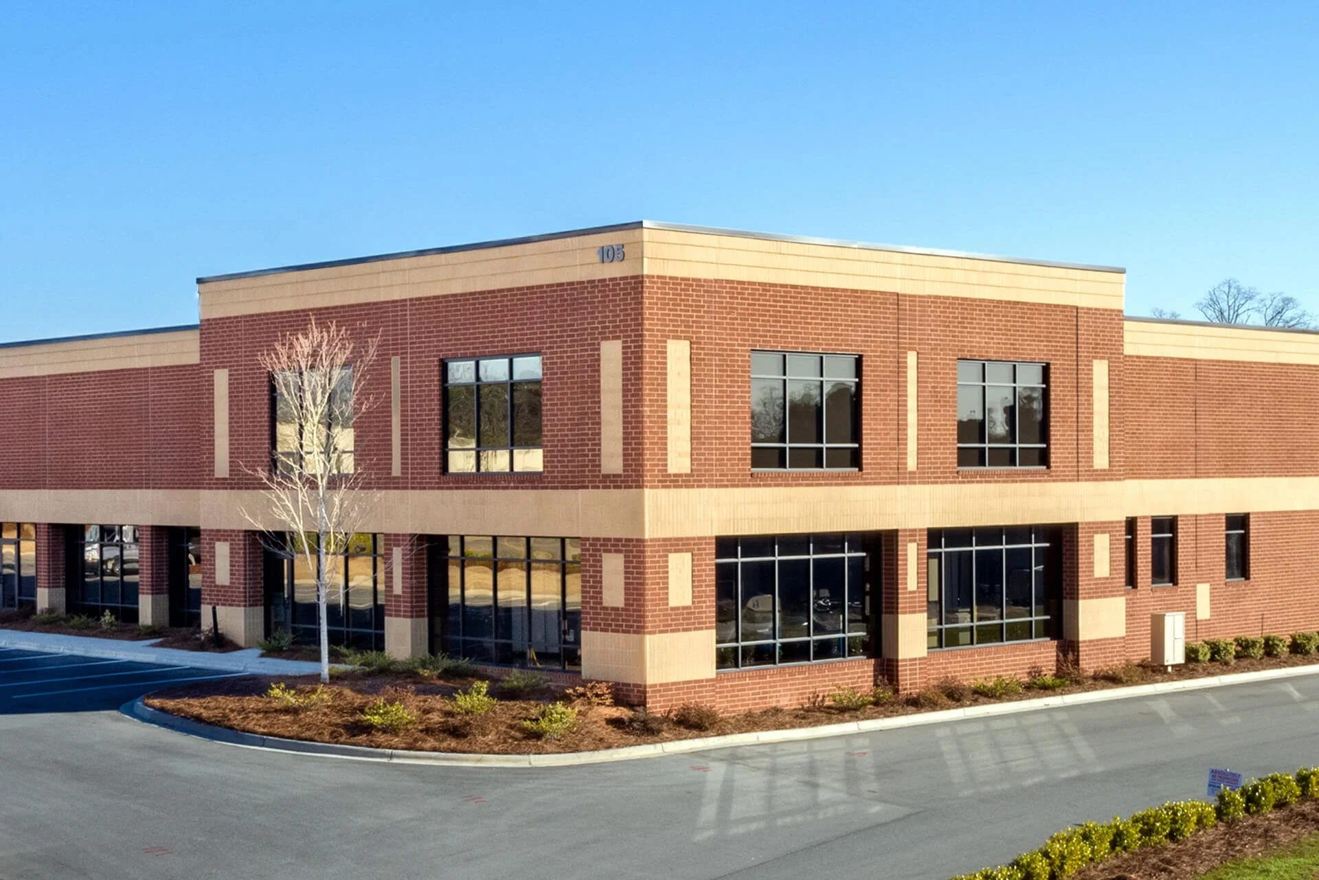 A two-story commercial building with red brick exterior and large glass windows, parking lot in front, and landscaped area with small bushes and a leafless tree.