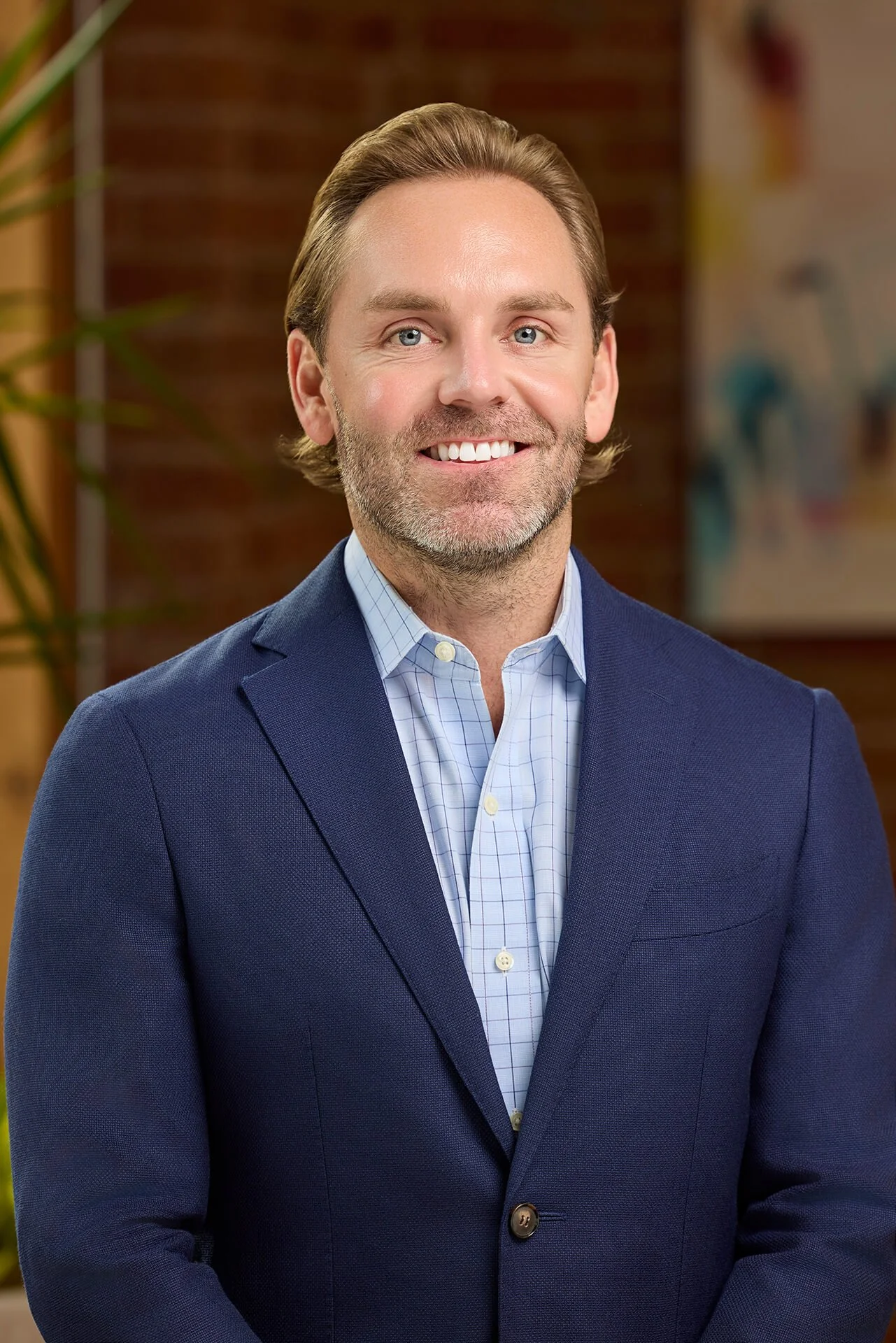 A smiling man in a blue suit and light blue checkered shirt posing indoors with a brick wall and blurred artwork in the background.