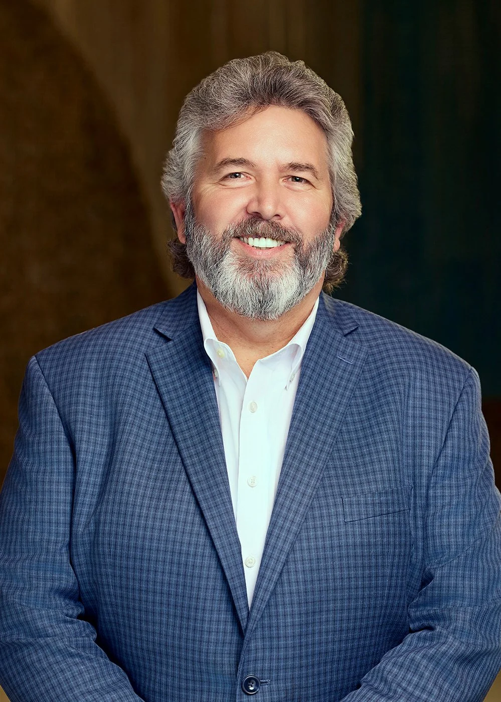 A middle-aged man with gray hair and a beard, wearing a blue checkered blazer and white shirt, smiling in a professional portrait.