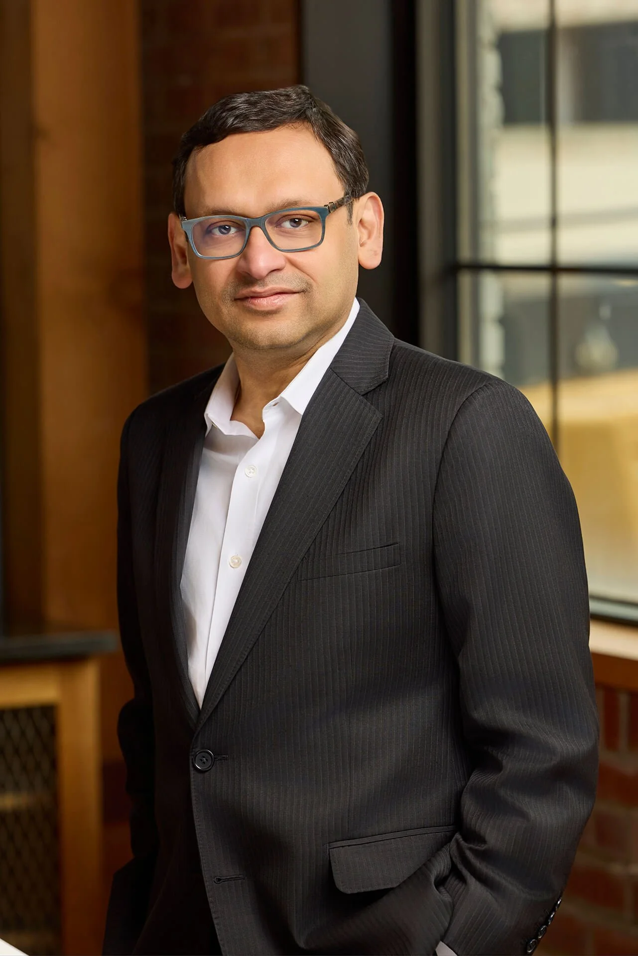 A man in a black suit and white shirt posing indoors near a large window.