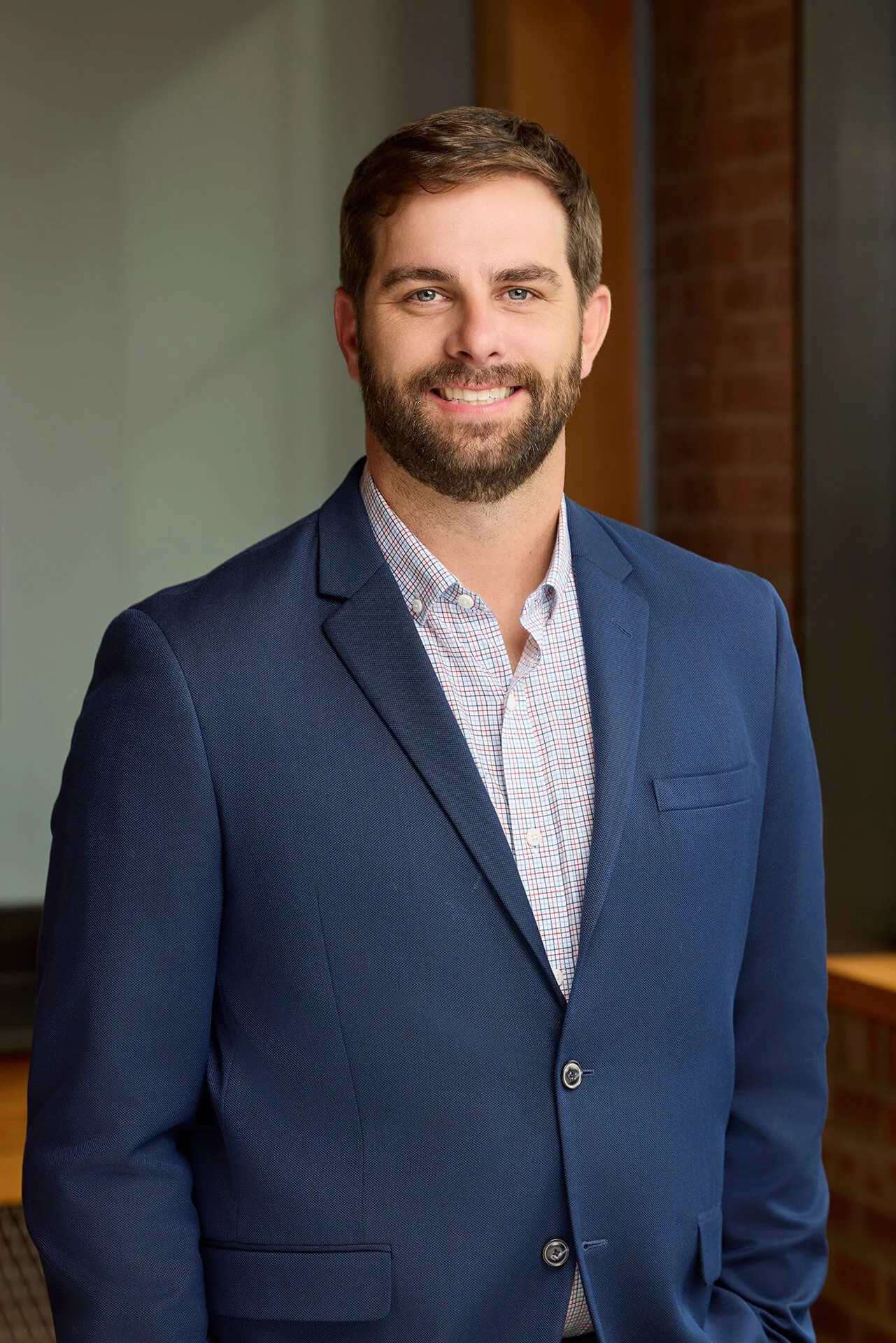 Professional man in a navy blue suit and checkered shirt standing indoors with a smile.