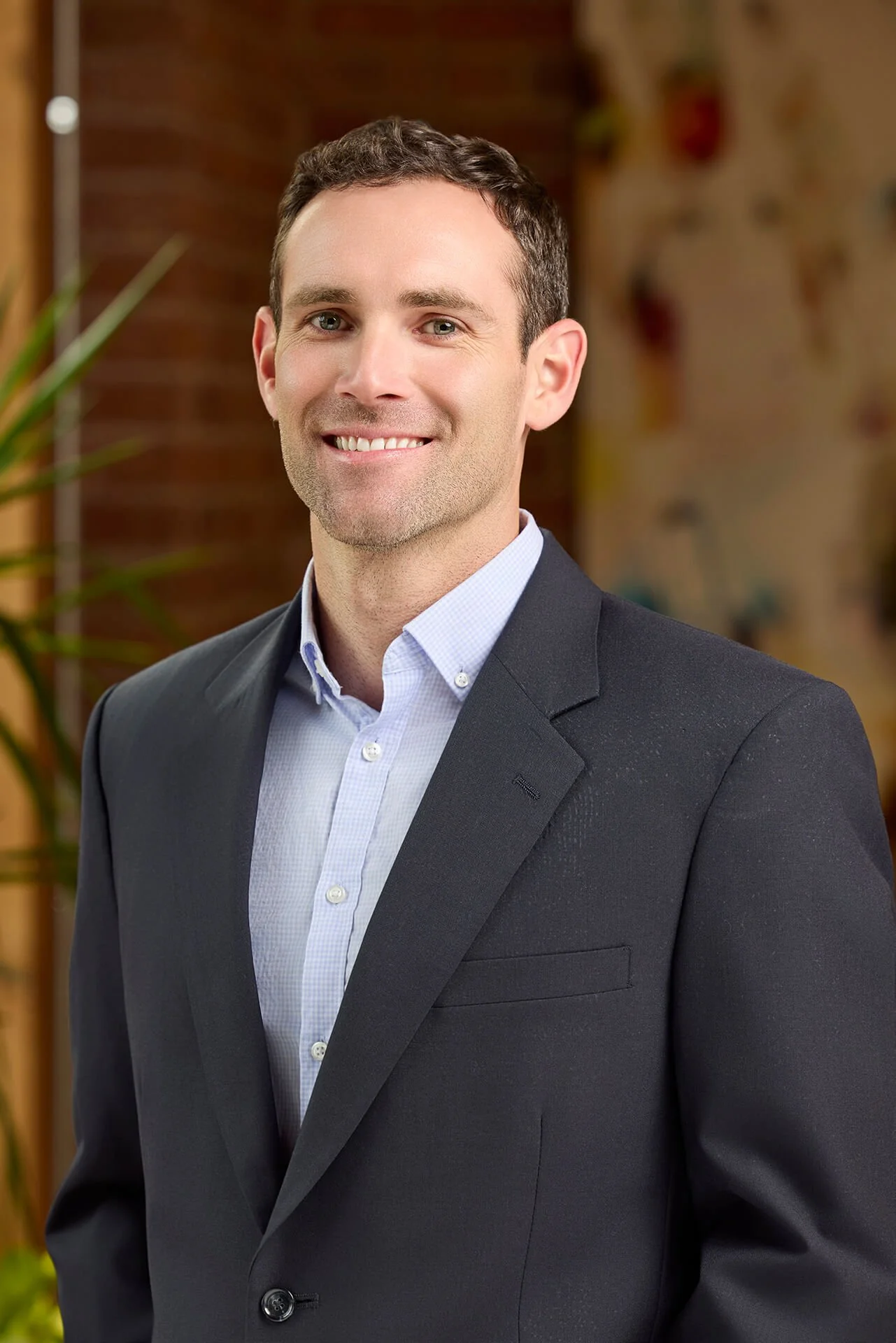 A man in a dark suit jacket and a light blue collared shirt, smiling, standing indoors with a brick wall background and some plants.
