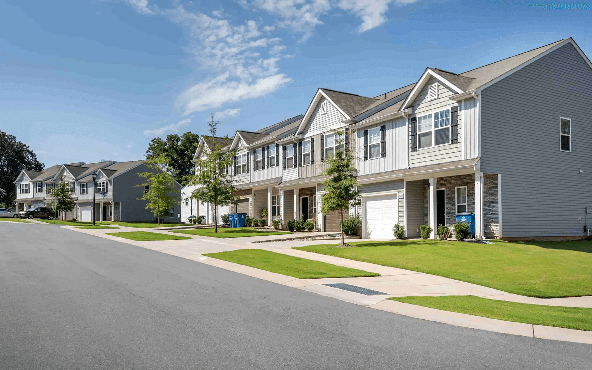A row of modern townhouses with front porches, small trees, and manicured lawns on a clean residential street under a blue sky with scattered clouds.