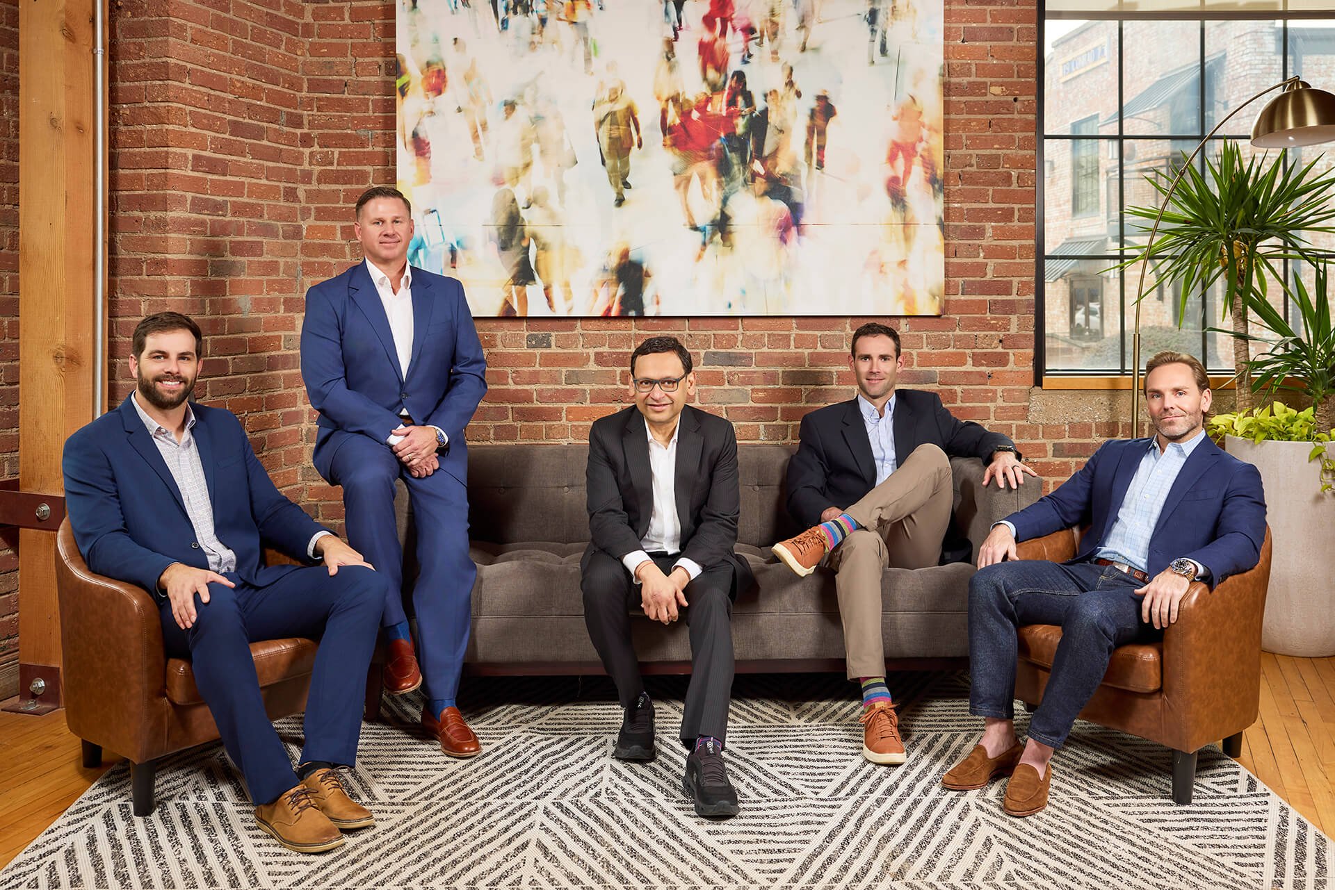 Five men in business attire posing in a modern office with exposed brick walls, a large colorful artwork, and a window with a cityscape view. They are seated and standing on and around a brown sofa and leather chairs.