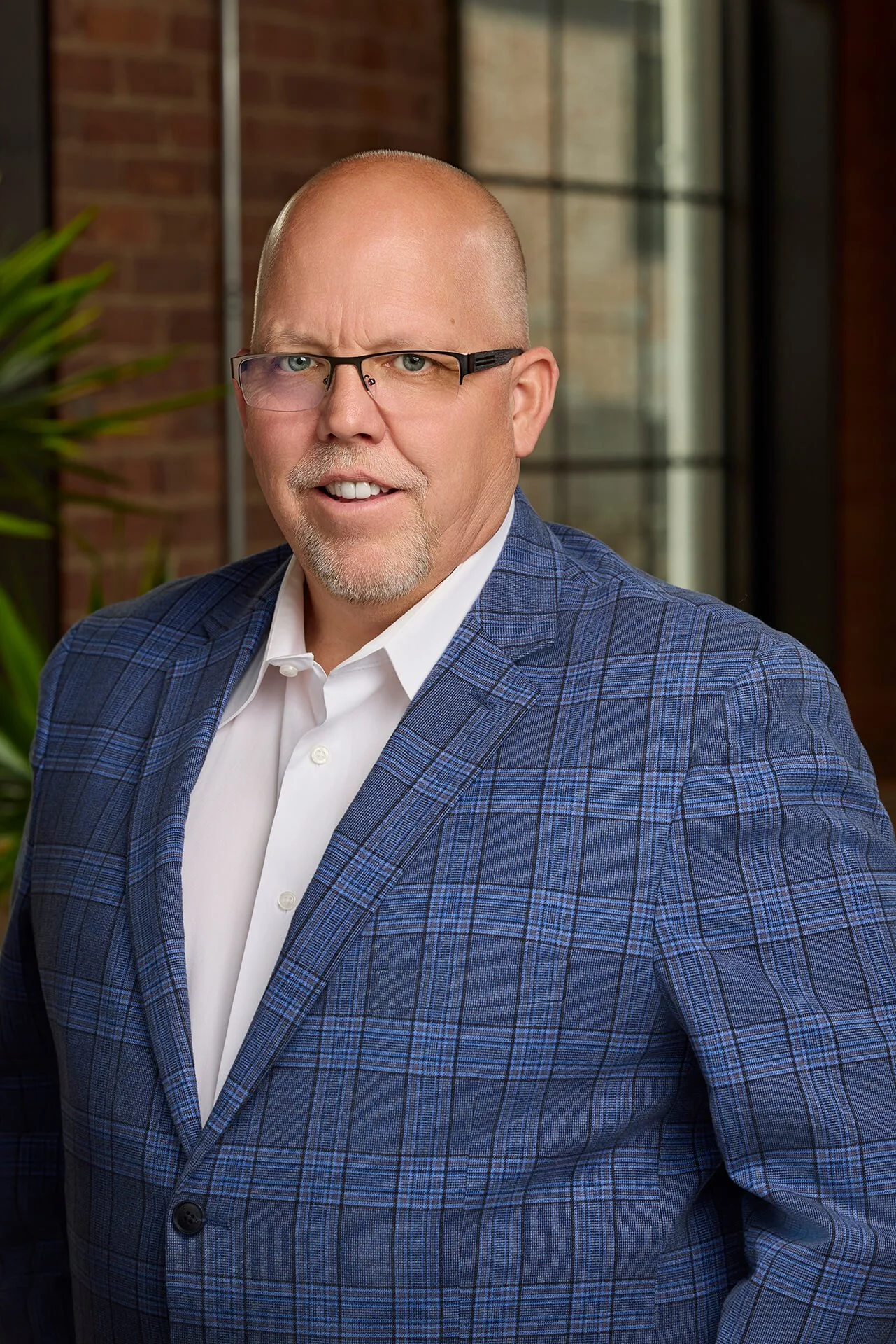 A middle-aged man with glasses, a bald head, and a goatee, dressed in a blue plaid jacket and white shirt, standing indoors with a brick wall, window, and green plant in the background.