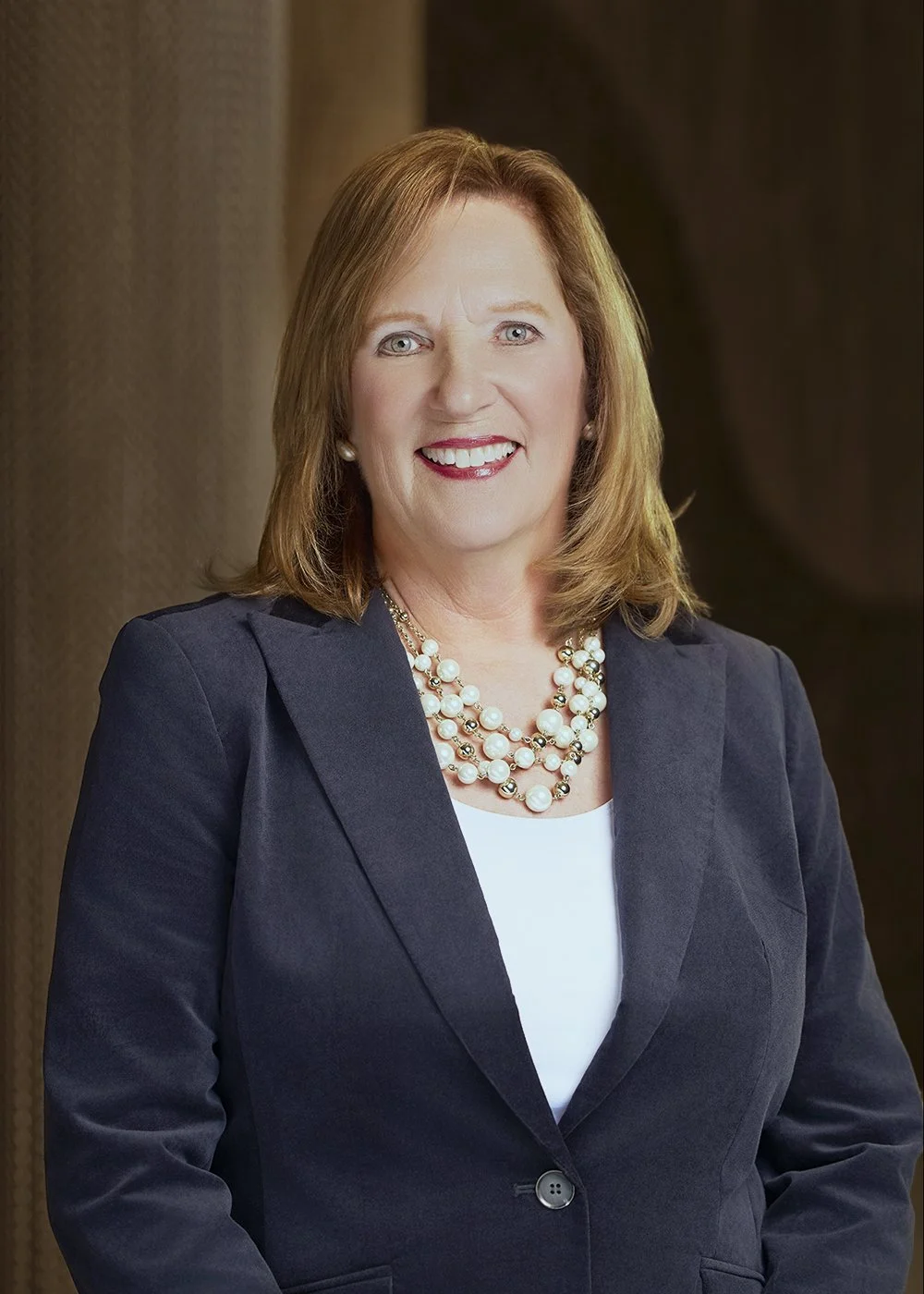 Professional woman with shoulder-length red hair, wearing a navy blazer, white top, and a layered pearl necklace, smiling in front of a dark background.