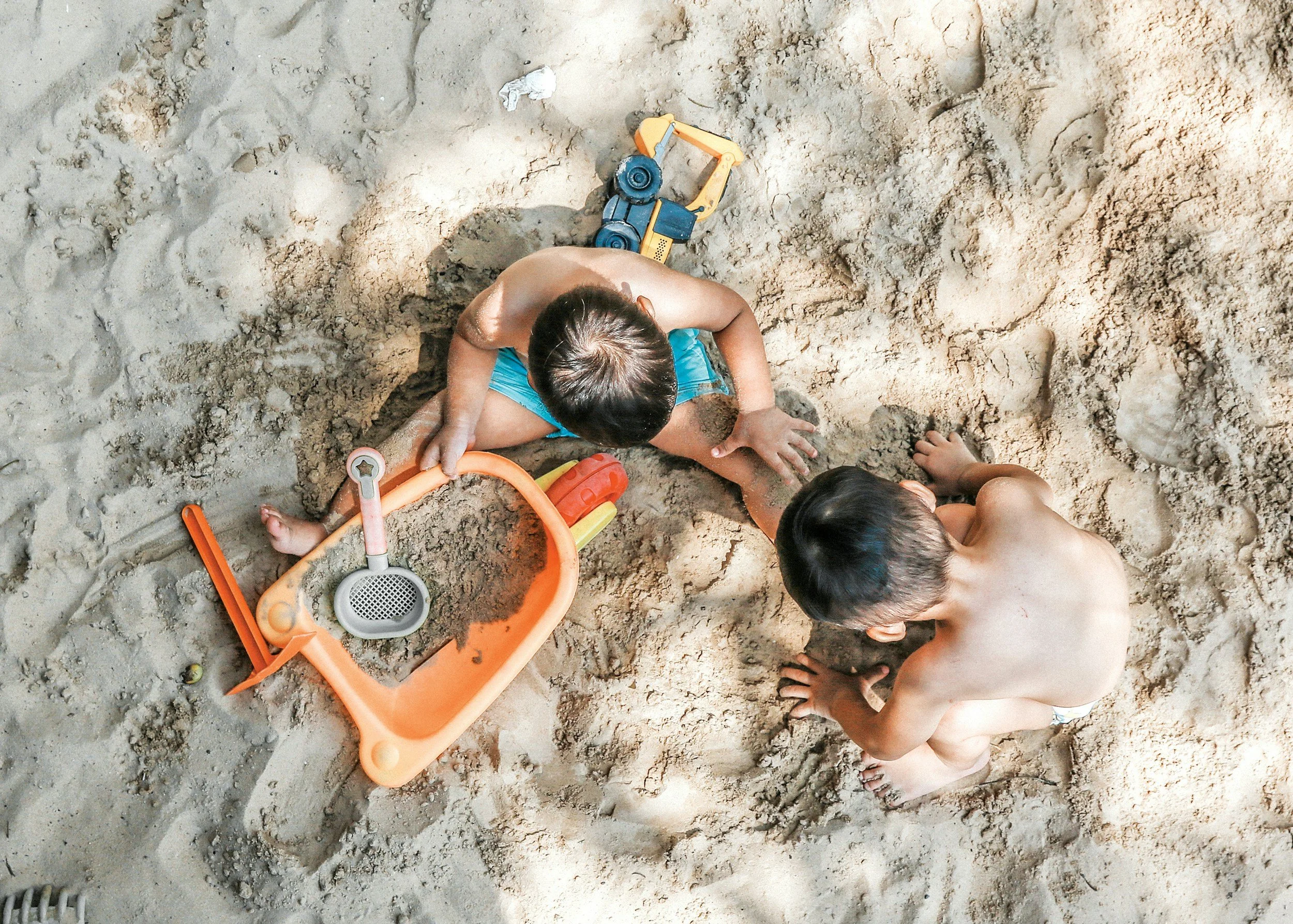Two young boys playing in the sand at the beach, with beach toys including a shovel, sand sieve, and an orange sand wheelbarrow.