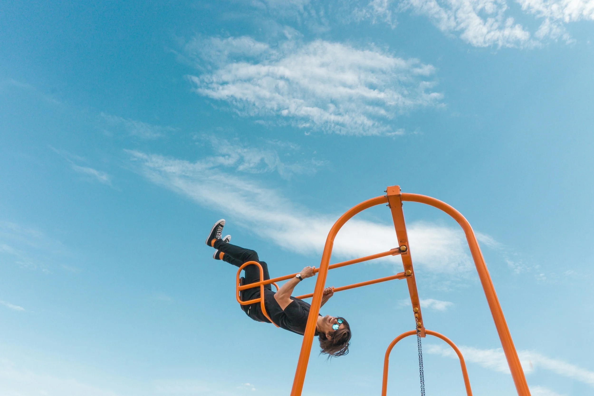 Person hanging upside down on orange playground equipment outdoors under a blue sky with clouds.
