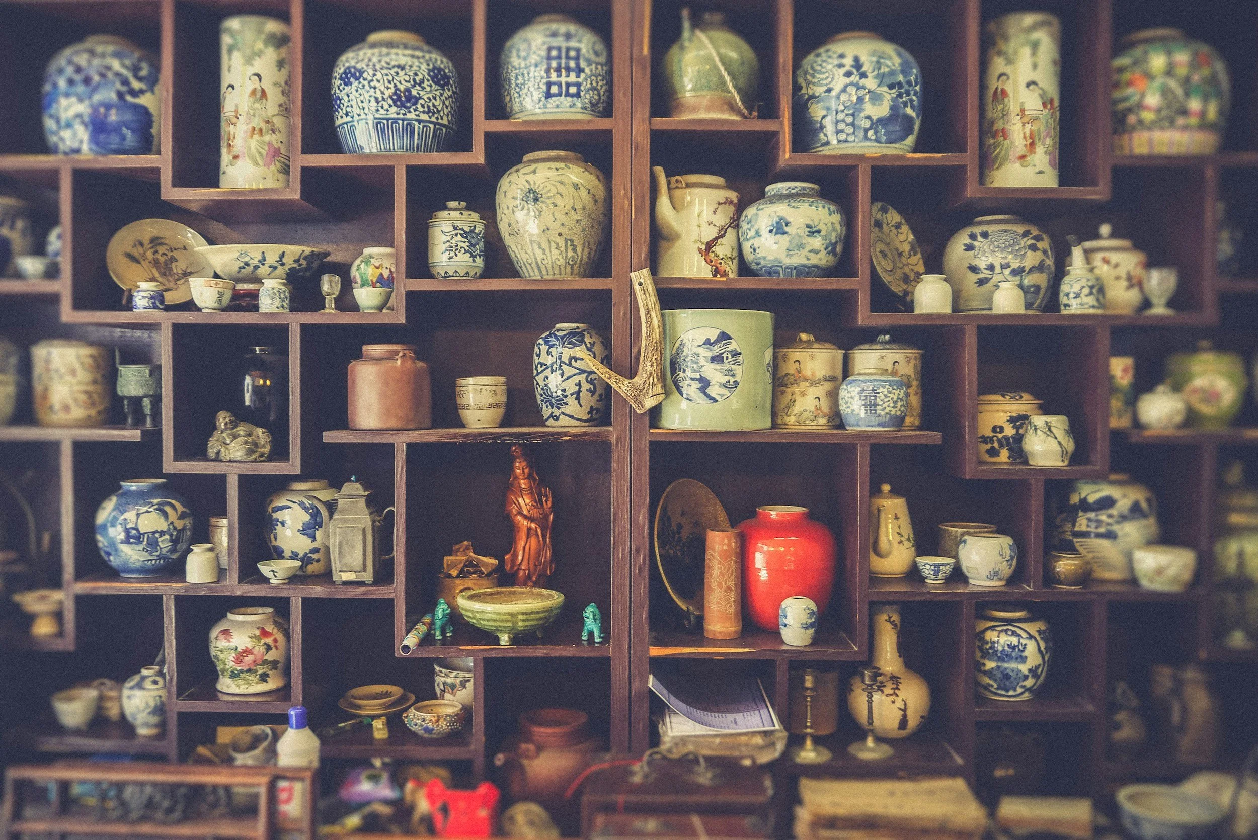 Shelf filled with various traditional Asian porcelain vases, jars, and decorative items, including figurines, a teapot, and small bowls, arranged in wooden cubbies.