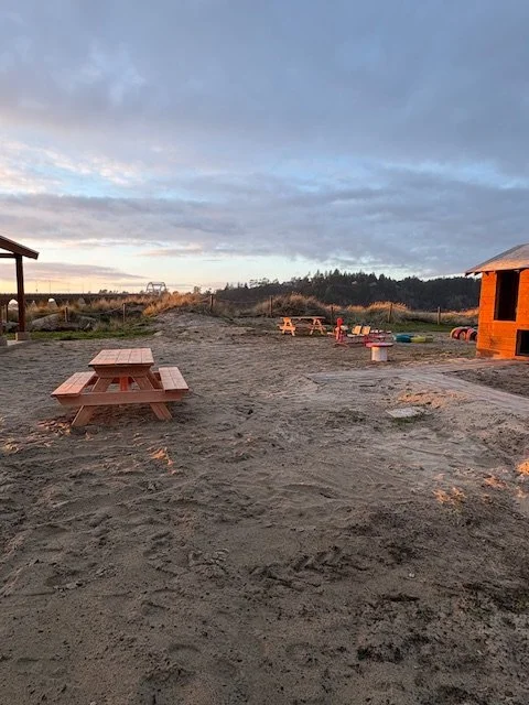 Picnic grounds with sand and picnic tables, the alsea bay bridge in the distance, and a sunset