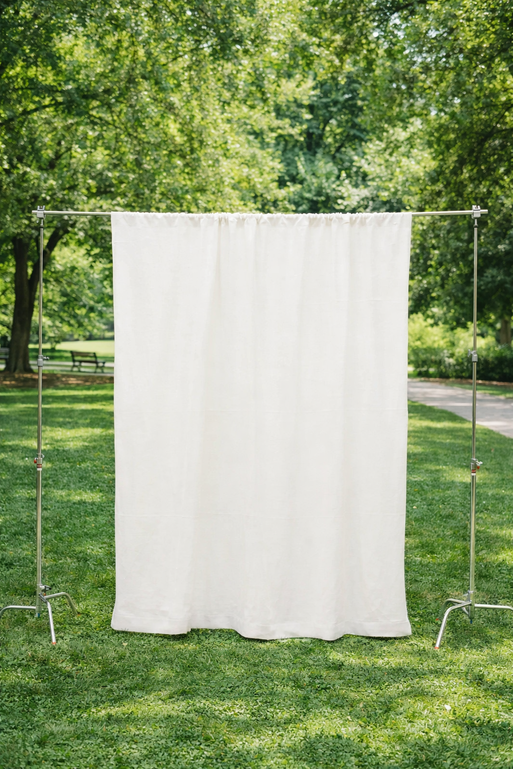 A white cloth backdrop hanging on a metal frame in a park with green trees and grass, benches, and a walking path in the background.