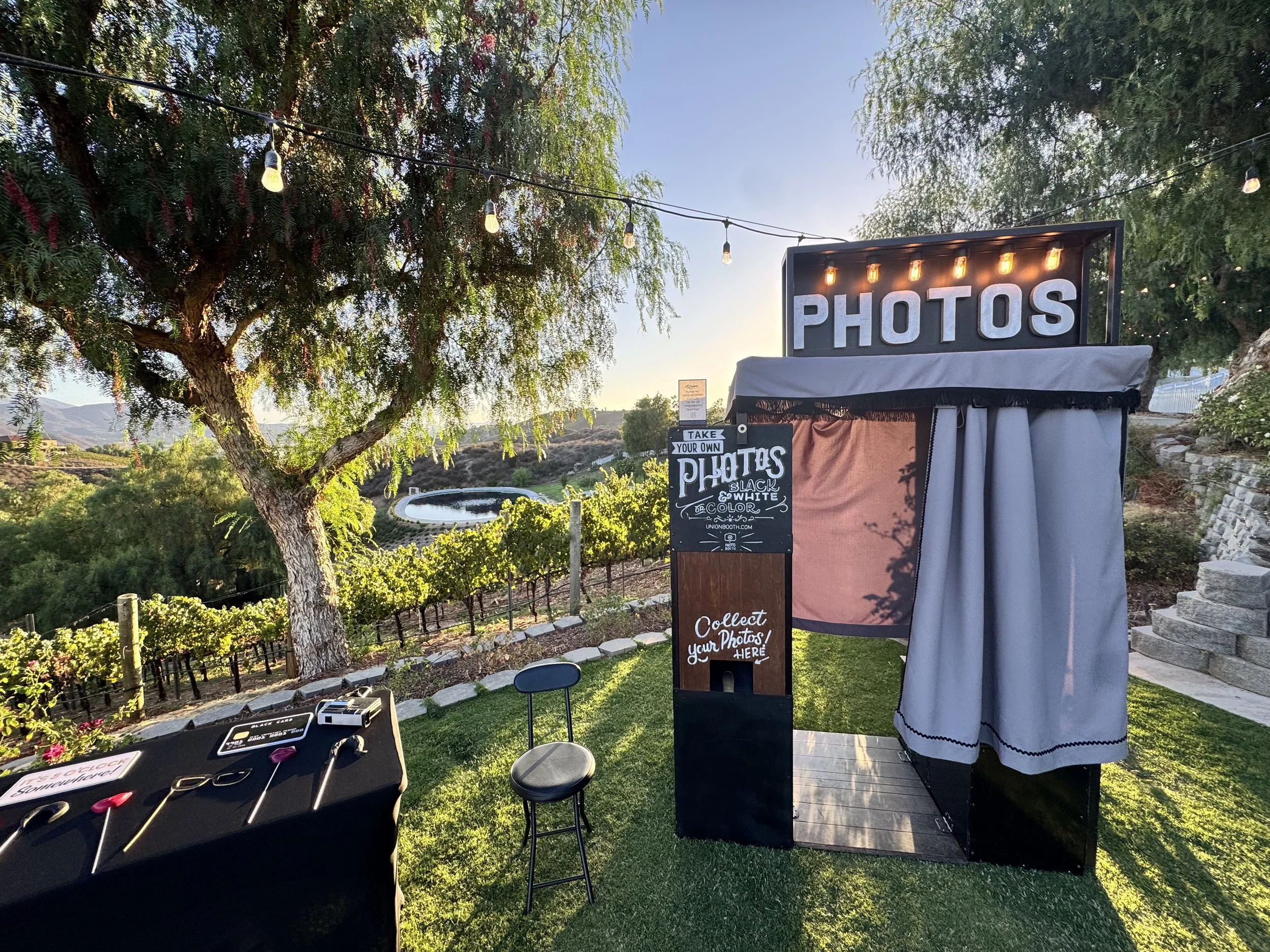 Photo booth setup with a large illuminated sign saying 'PHOTOS', a small chalkboard sign inviting to take or collect photos, a black stool, and a table with photo props, outdoors near trees, vineyards, and a swimming pool, during sunset