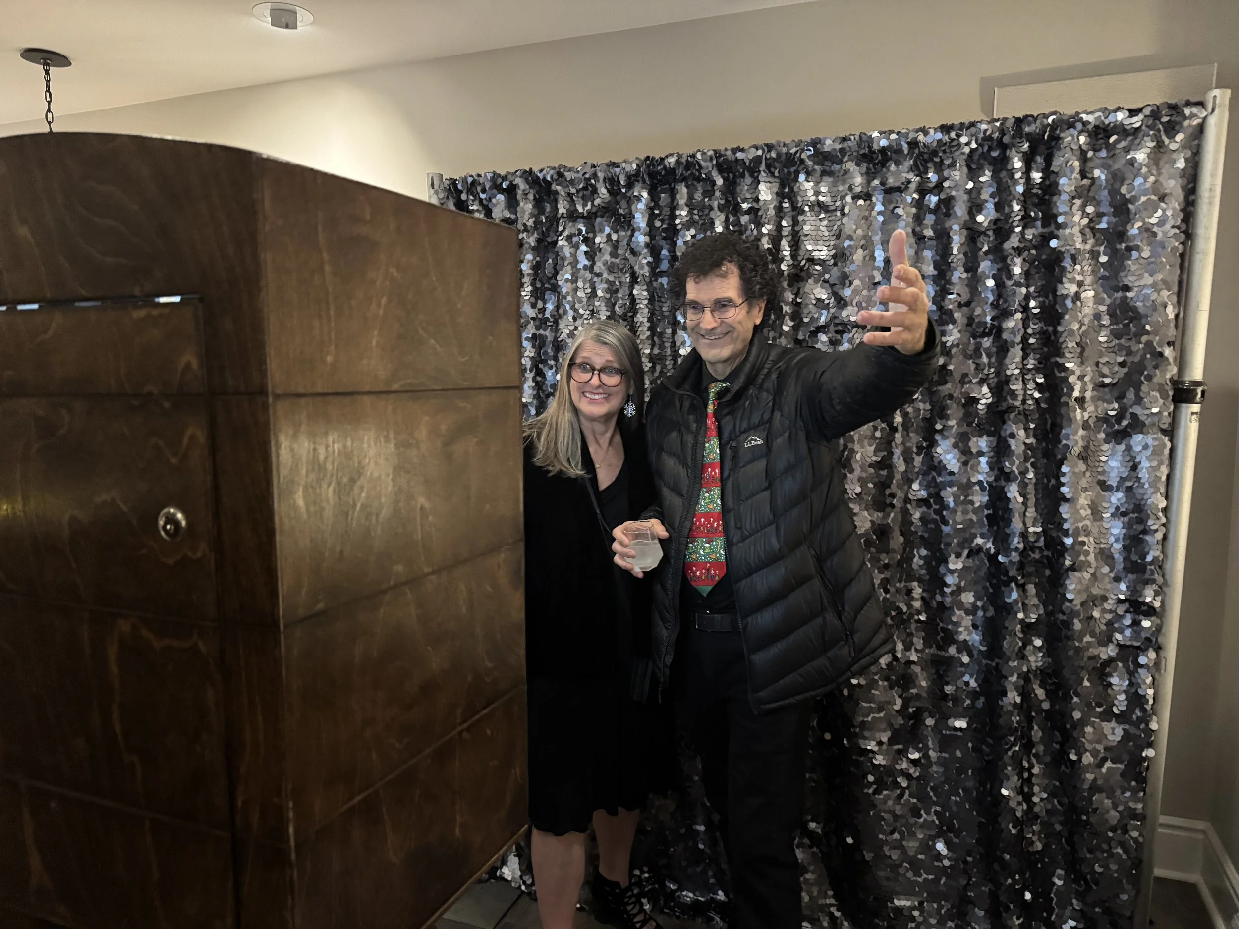 Two people, a woman and a man, are posing for a photo in front of a black and silver glittery backdrop. The woman has blonde hair, glasses, and is wearing a black dress. The man has curly hair, glasses, and is wearing a black puffy jacket with a festive Christmas tie. The man is smiling and gesturing with his right hand, while holding a drink in his left hand.