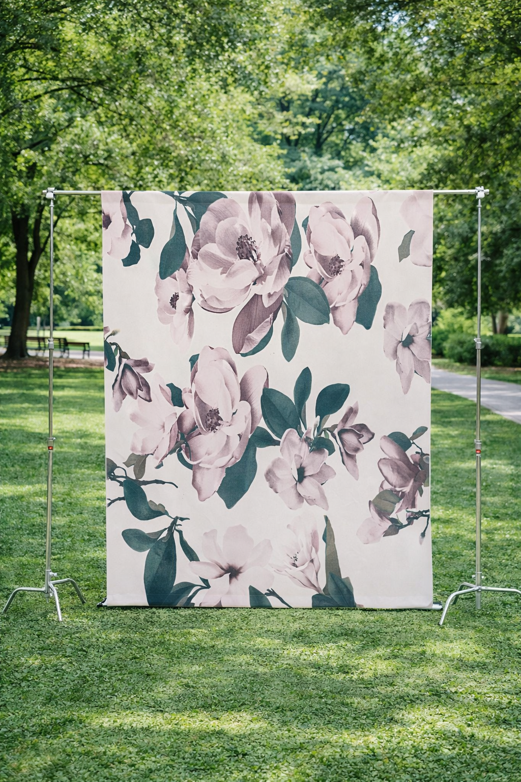 A floral fabric with pink and purple flowers and green leaves hanging on a metal frame outdoors in a park with green trees and grass.