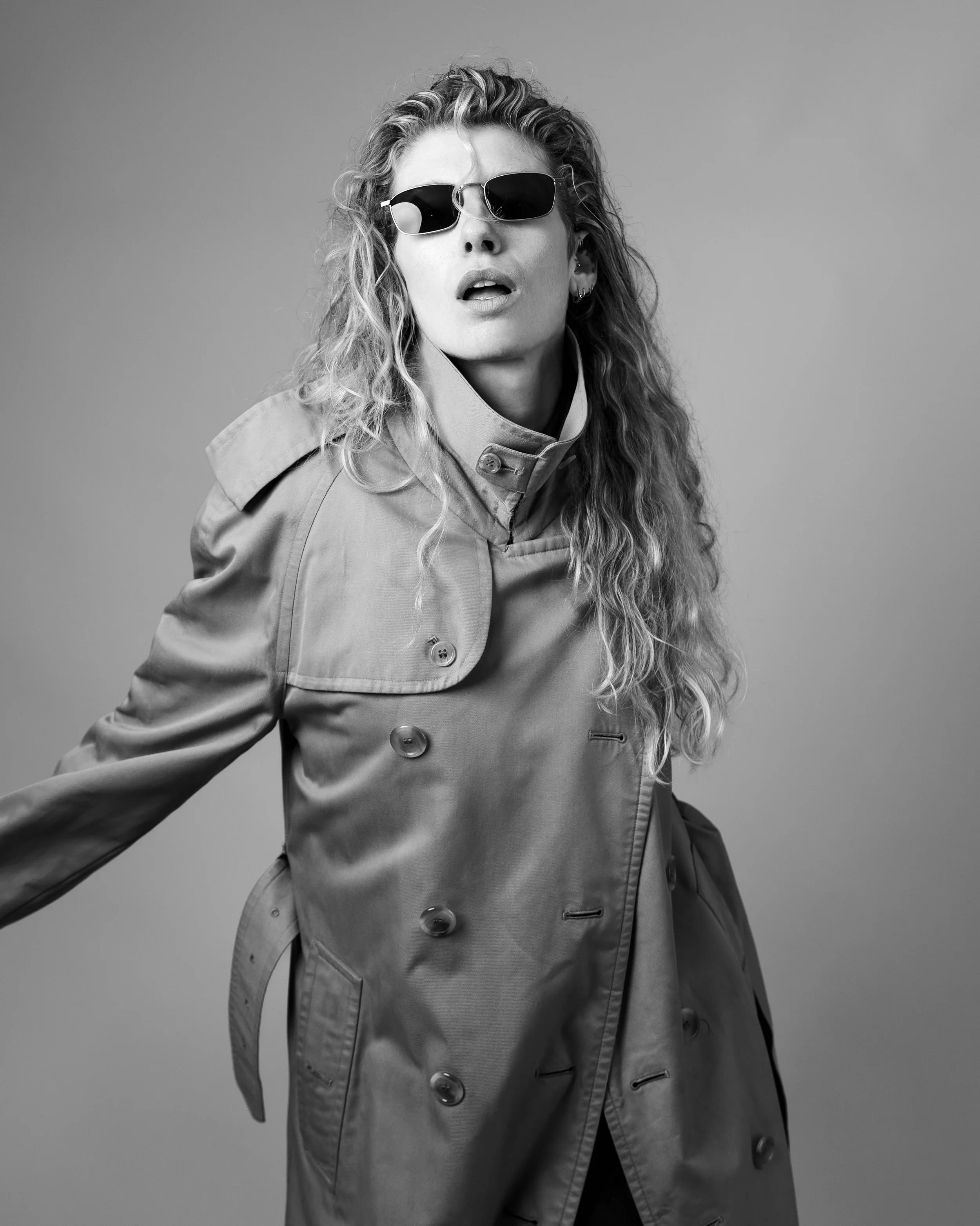 Black-and-white fashion portrait of a model wearing sunglasses and a high-collared trench coat in a London studio.